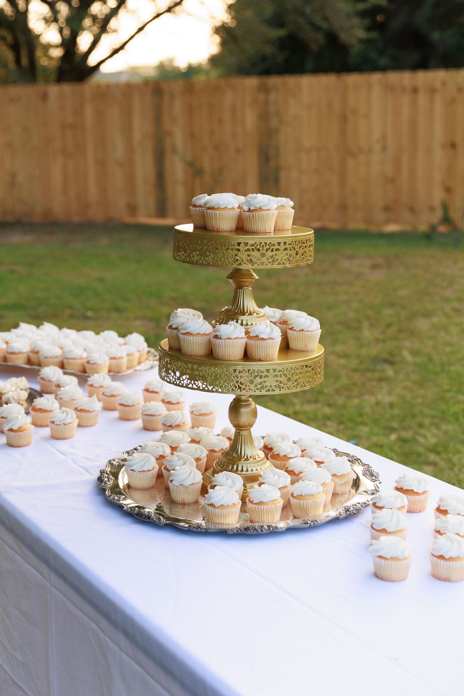 Elegant display of mini cupcakes arranged on white tablecloth with gold tiered serving stands at an outdoor event.