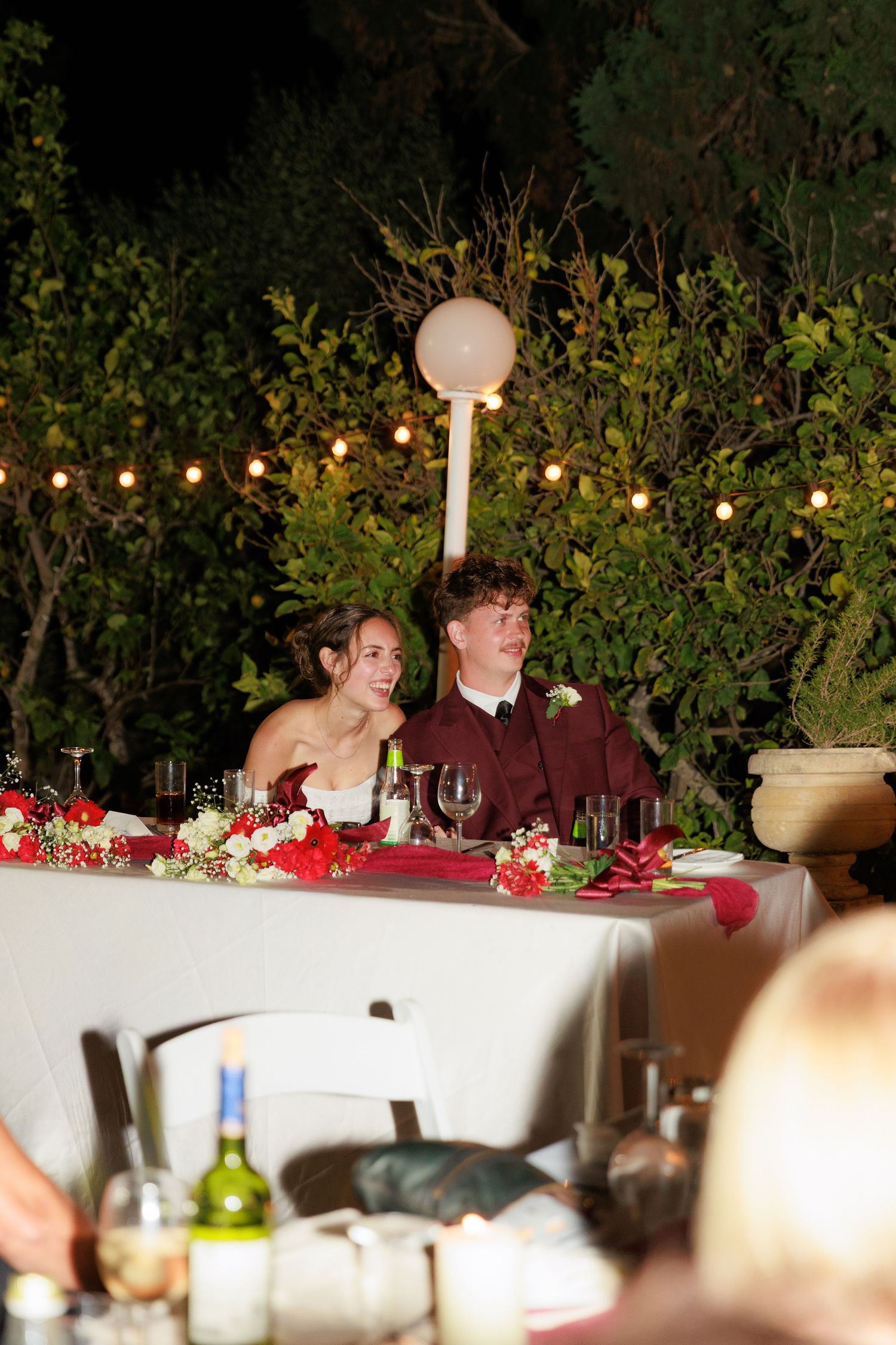 Elegant table setting at an outdoor evening reception with string lights and red floral centerpieces.