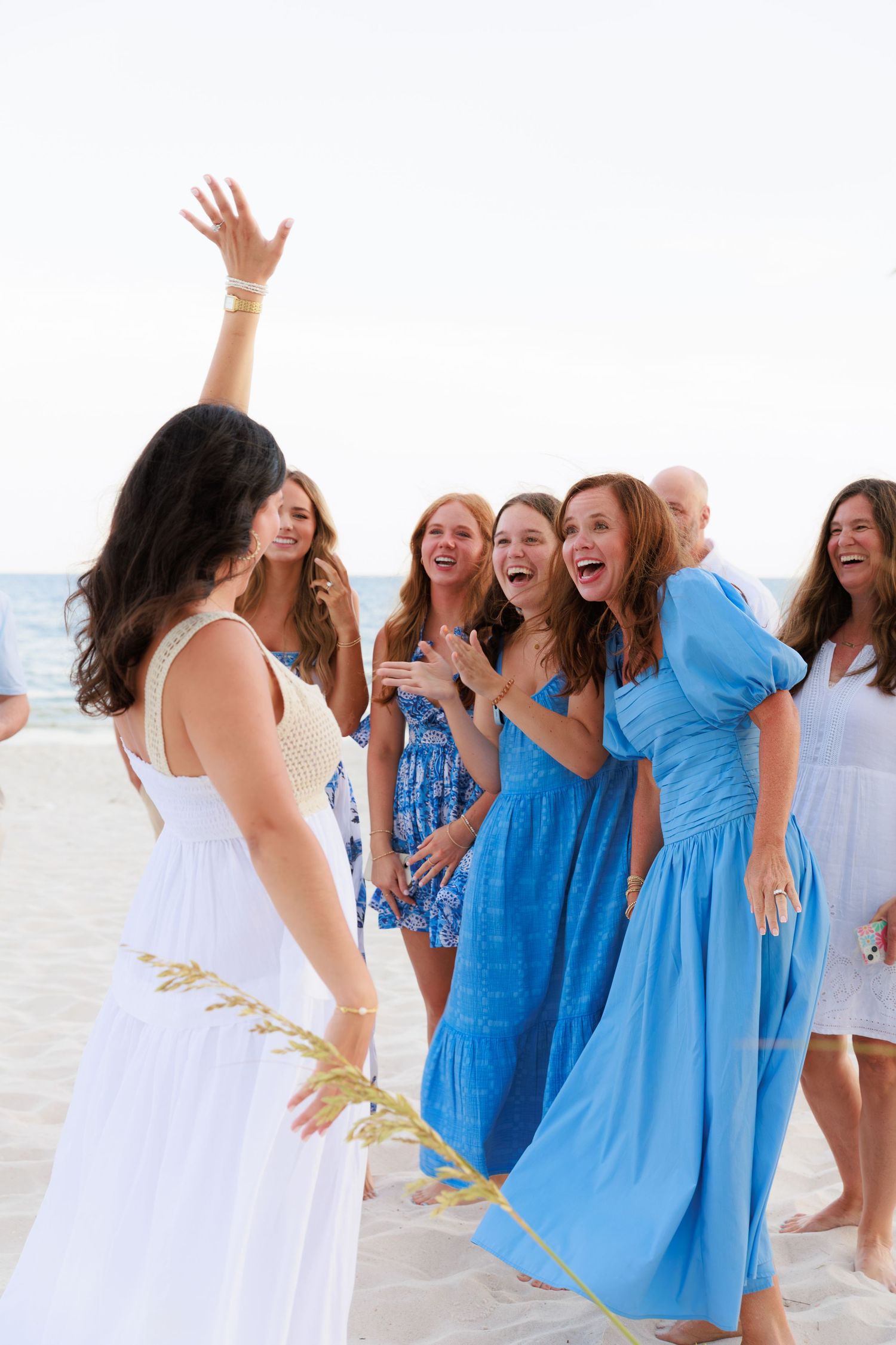 A group of bridesmaids in blue and white dresses celebrate joyfully on a sunny beach.