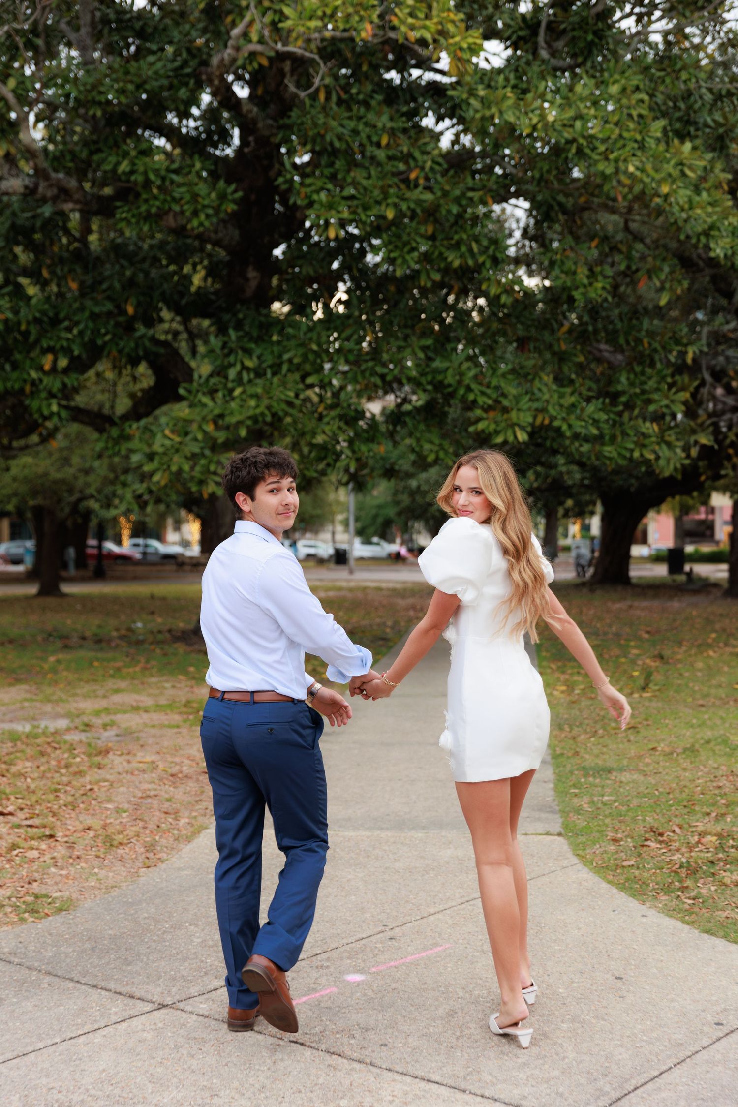 A playful couple walking and holding hands on a tree-lined pathway.