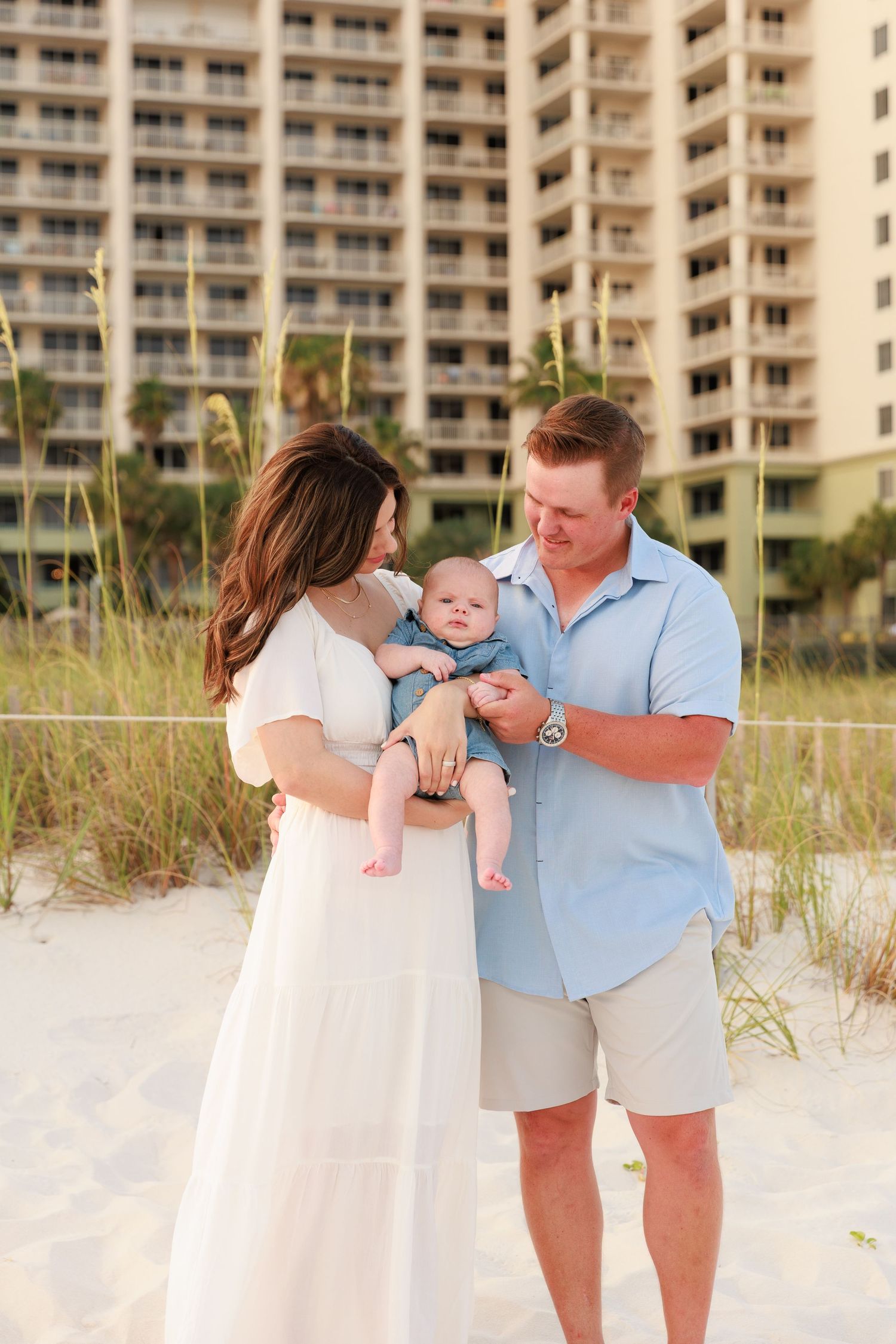 Young family with baby poses on sandy beach with coastal high-rise building backdrop.