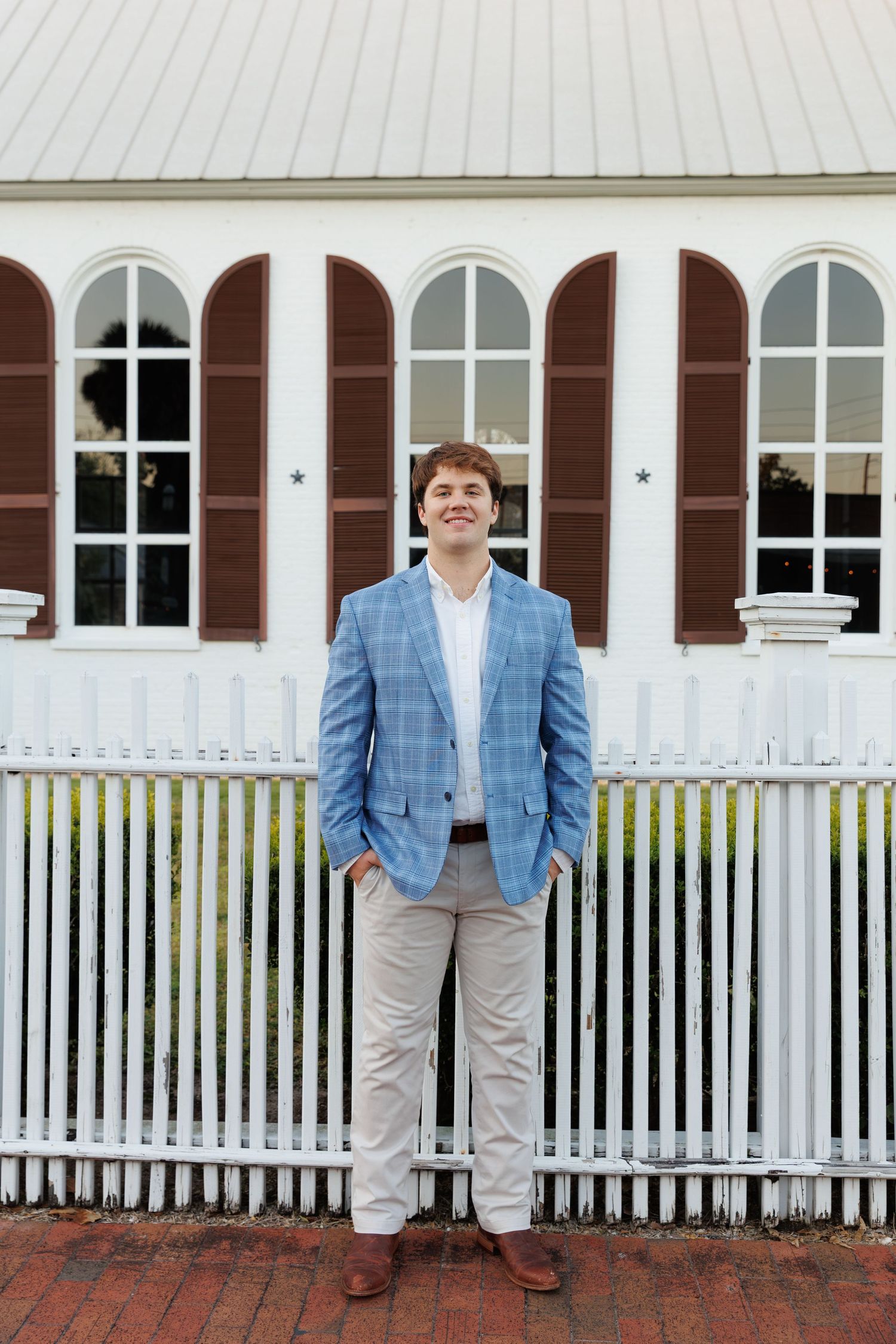 Young man in blue plaid blazer and khakis stands confidently by white picket fence outdoors.