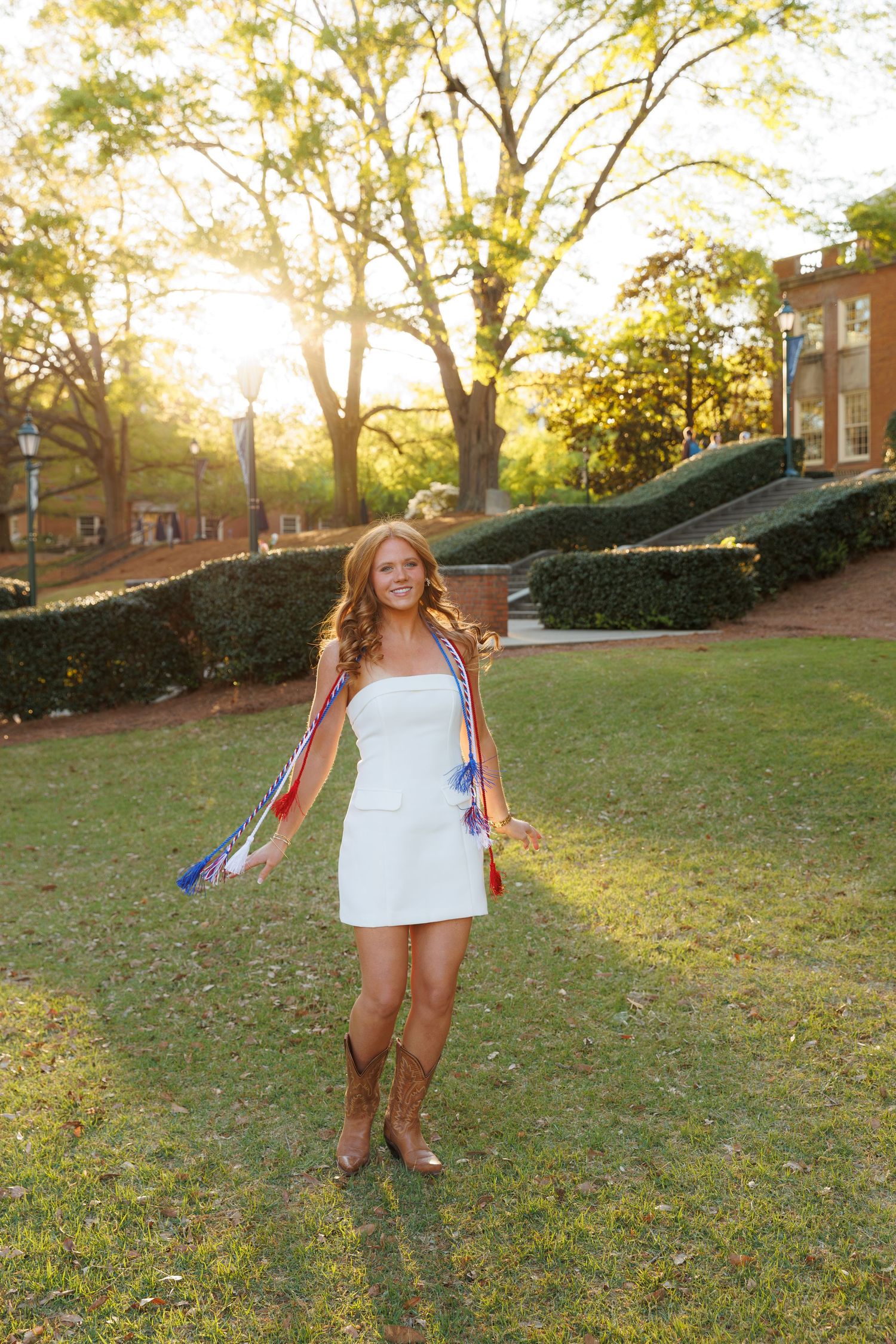Young woman in white dress and cowboy boots holding colorful sashes outdoors at golden hour.