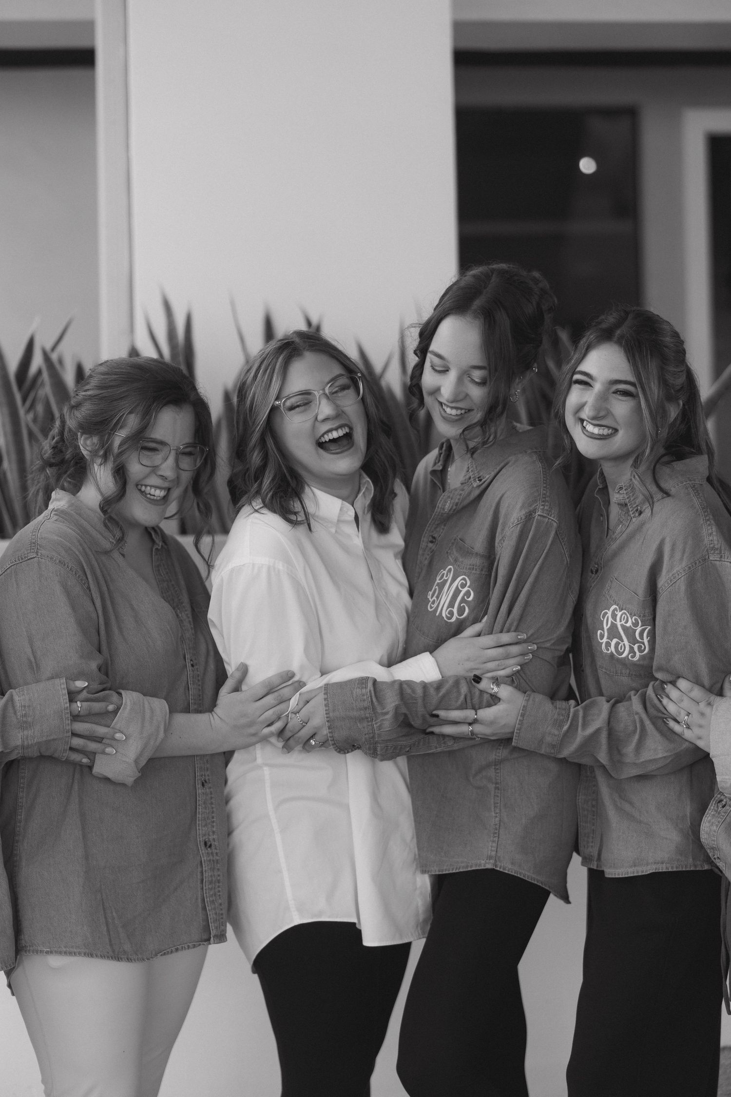 Four bridesmaids laughing together in monogrammed button-down shirts during wedding preparations in black and white photo.