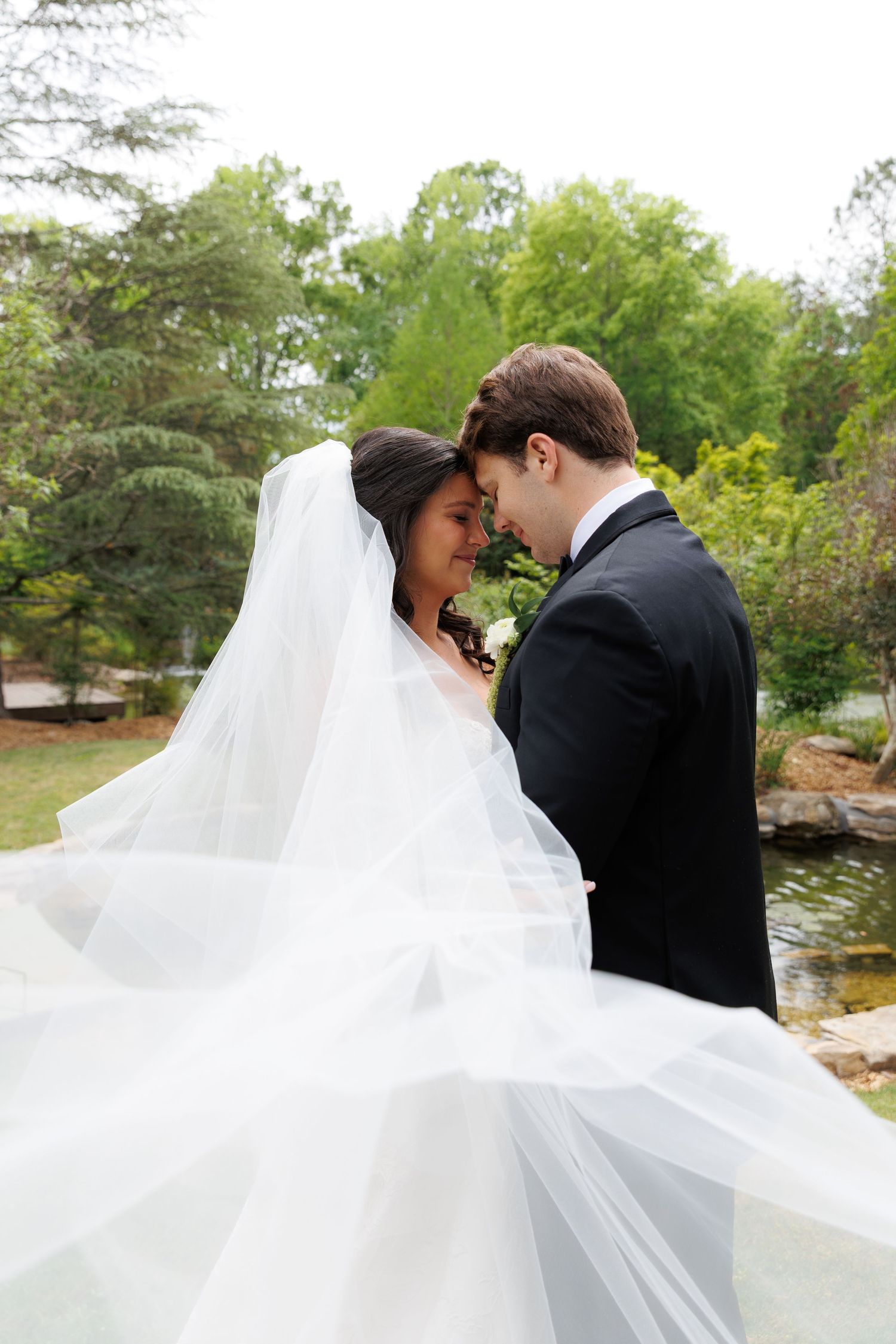 Bride and groom touching foreheads romantically outdoors, her veil flowing gracefully, surrounded by lush green trees near a pond.