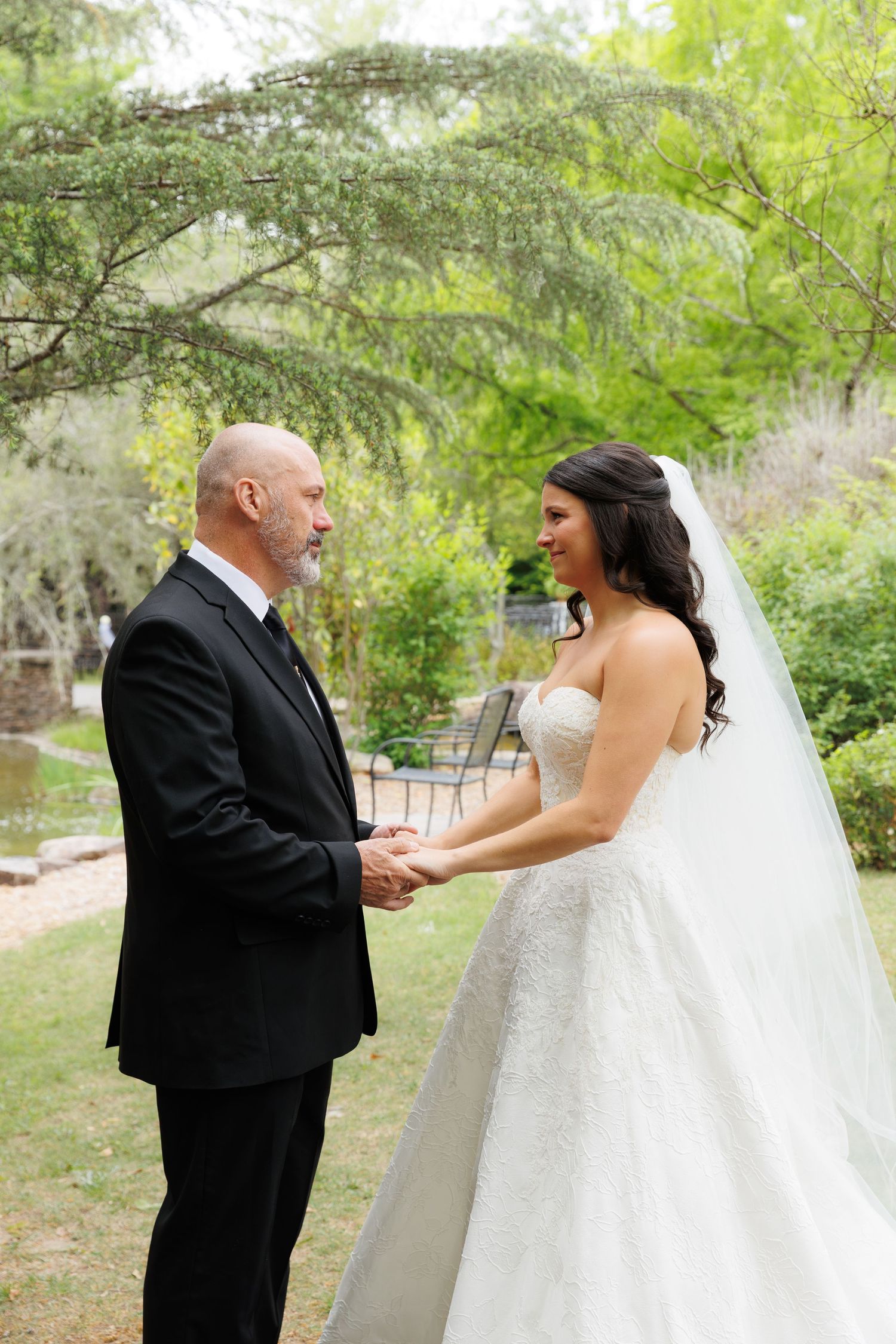 Bride and groom hold hands outdoors in a garden setting, bride wearing a strapless lace gown with veil.