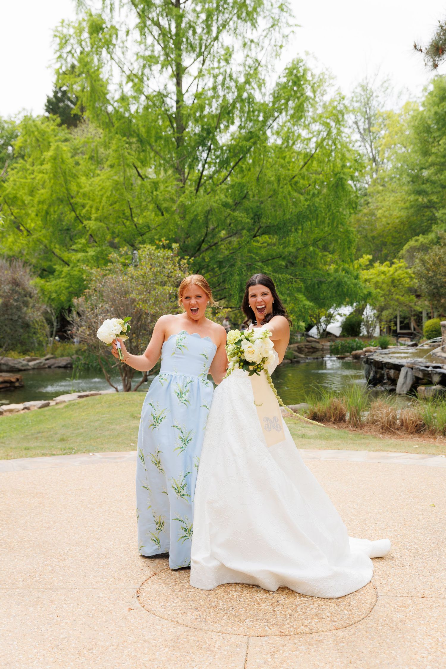 Bride in white gown and bridesmaid in blue floral dress holding bouquets, posing in a garden with pond.