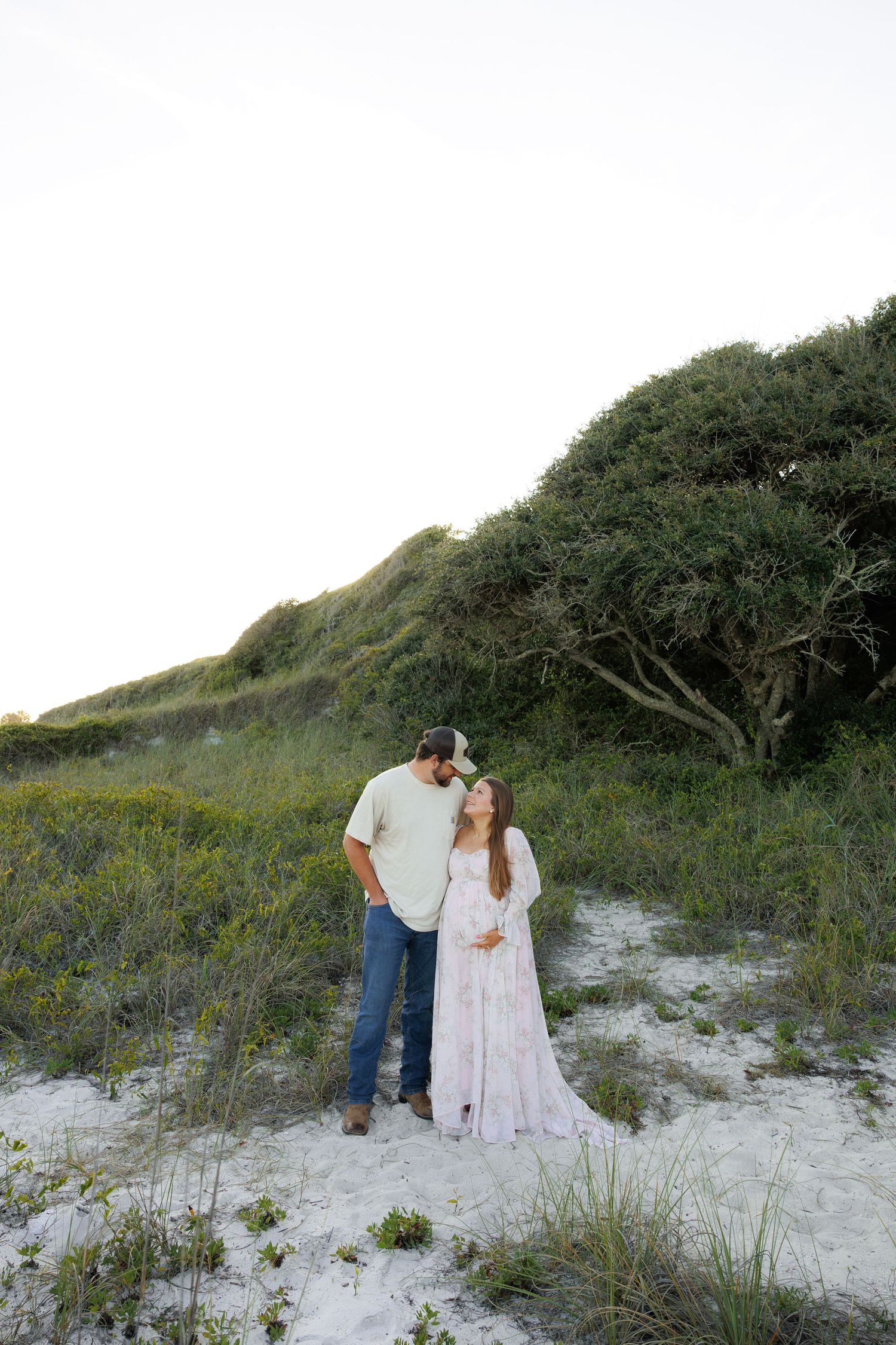 Couple posing on sandy beach dunes with lush greenery, woman in floral maternity gown, man in jeans and white shirt.