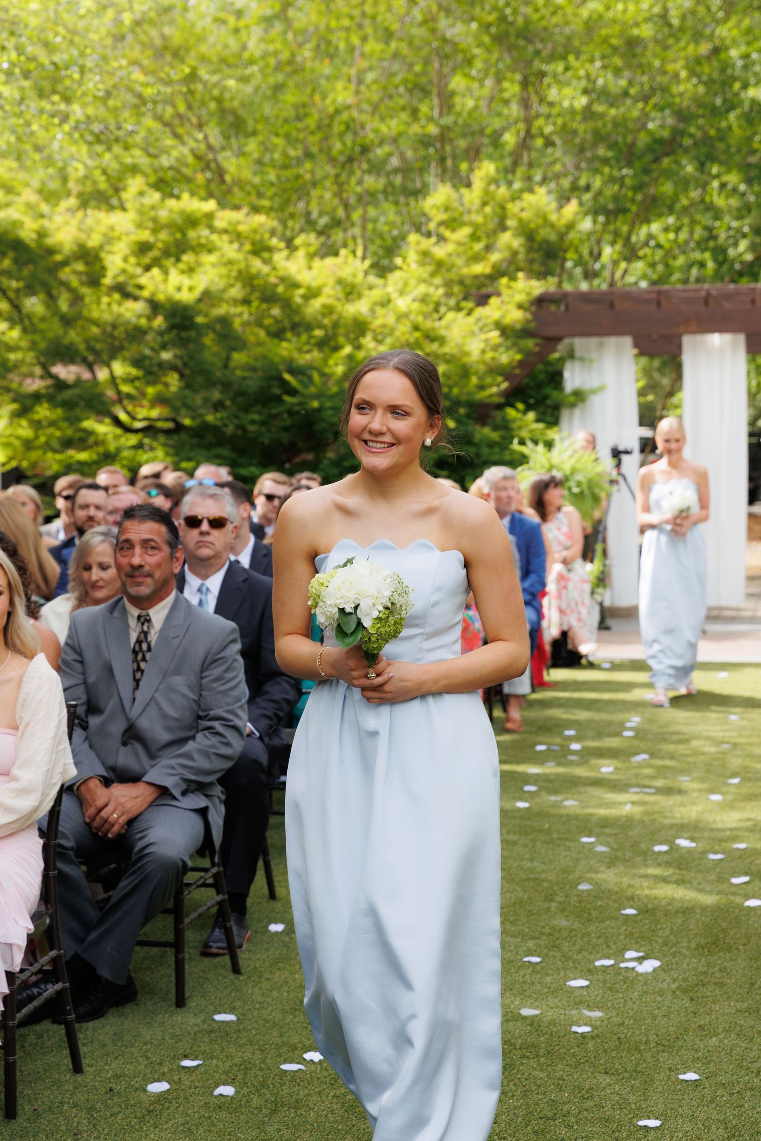Bridesmaid in light blue strapless gown carries white bouquet down outdoor wedding aisle.