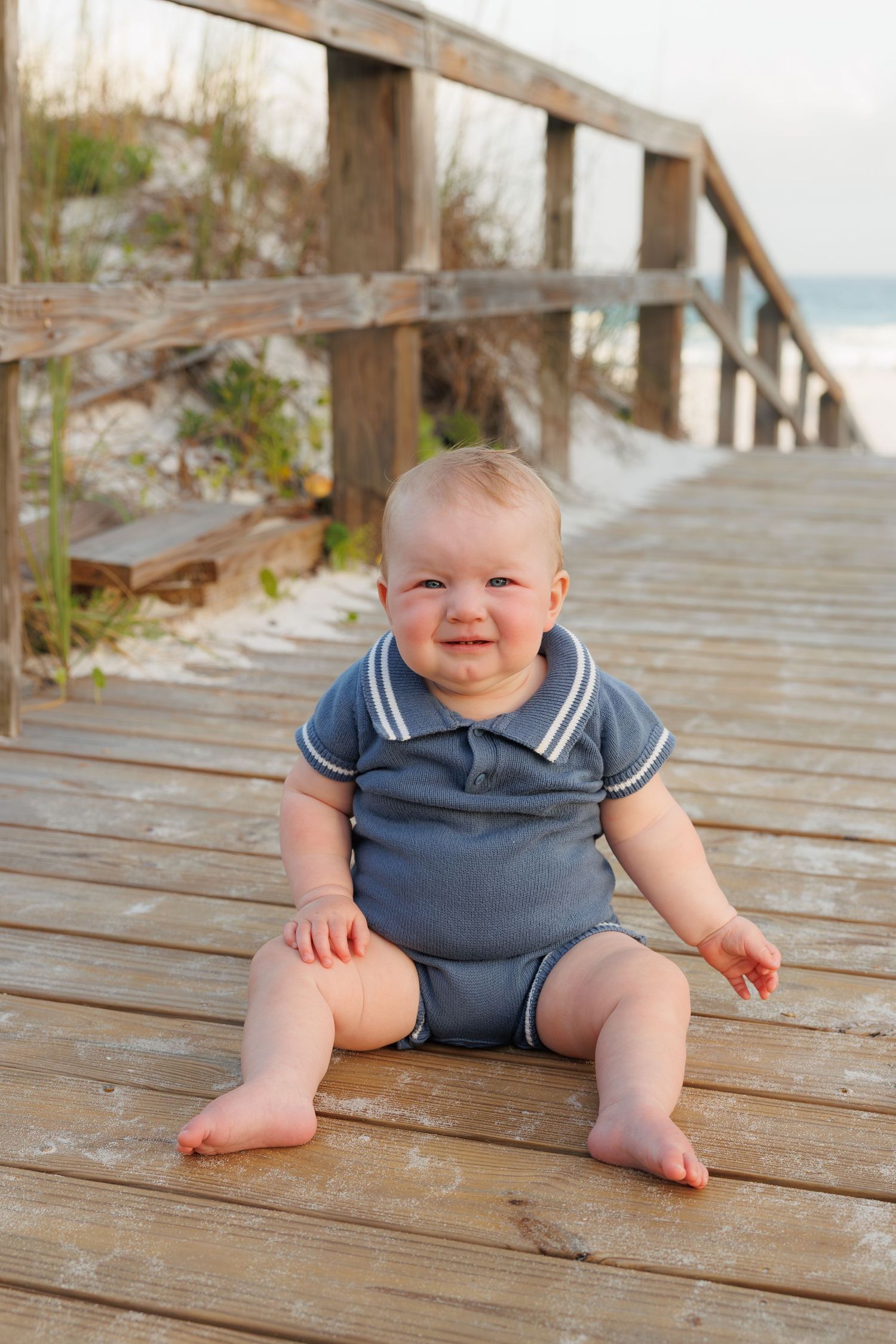 Adorable baby boy in blue romper sitting on a wooden beach boardwalk.