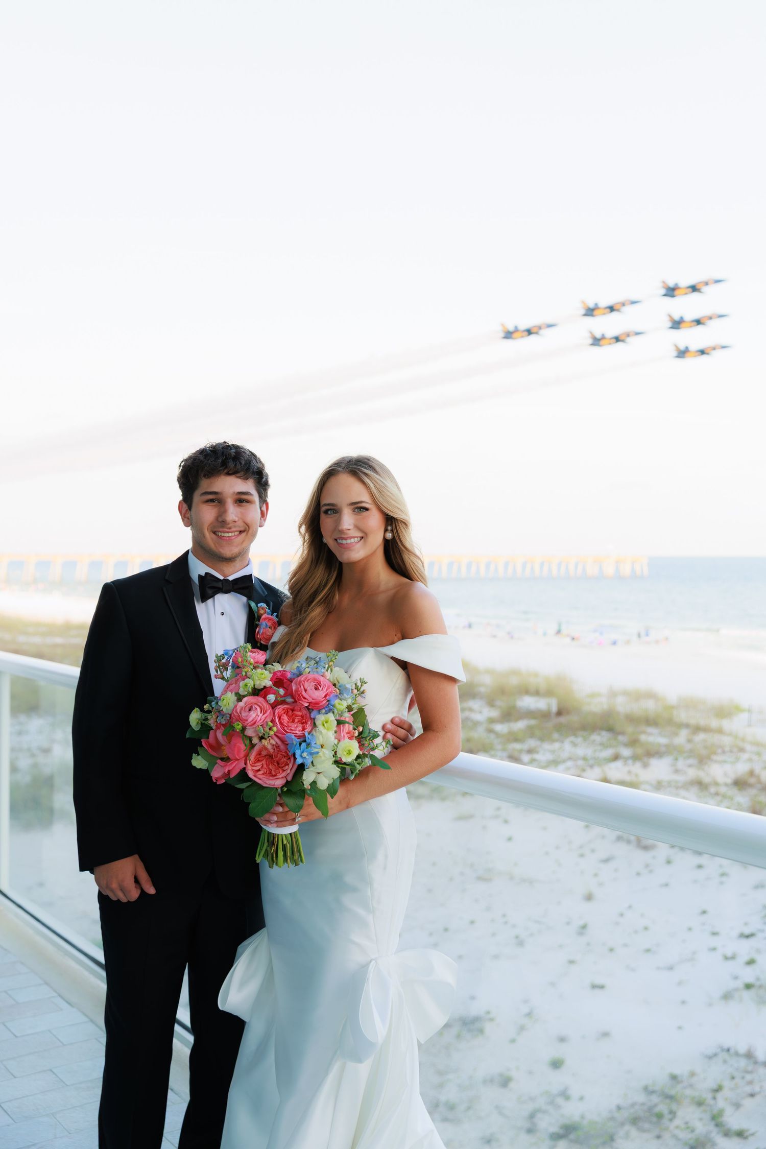 A beach wedding couple poses together on a balcony overlooking the ocean while holding a vibrant bouquet.