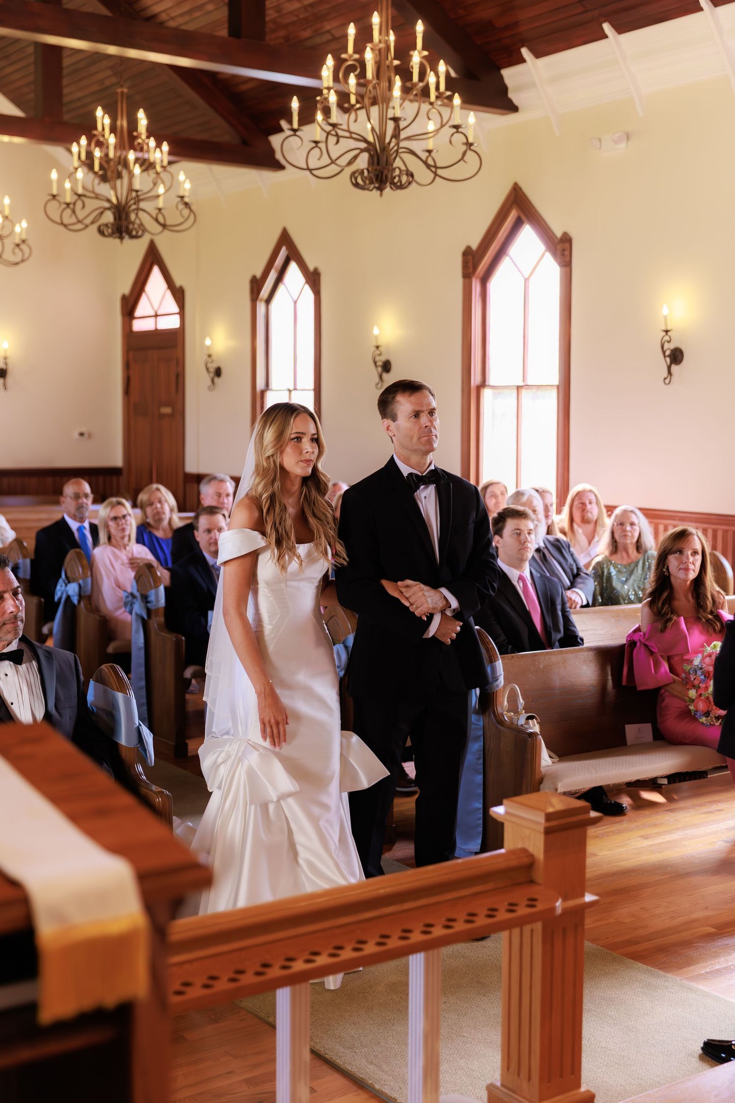 Inside a chapel with chandeliers and arched windows, a wedding ceremony takes place with guests seated in wooden pews.