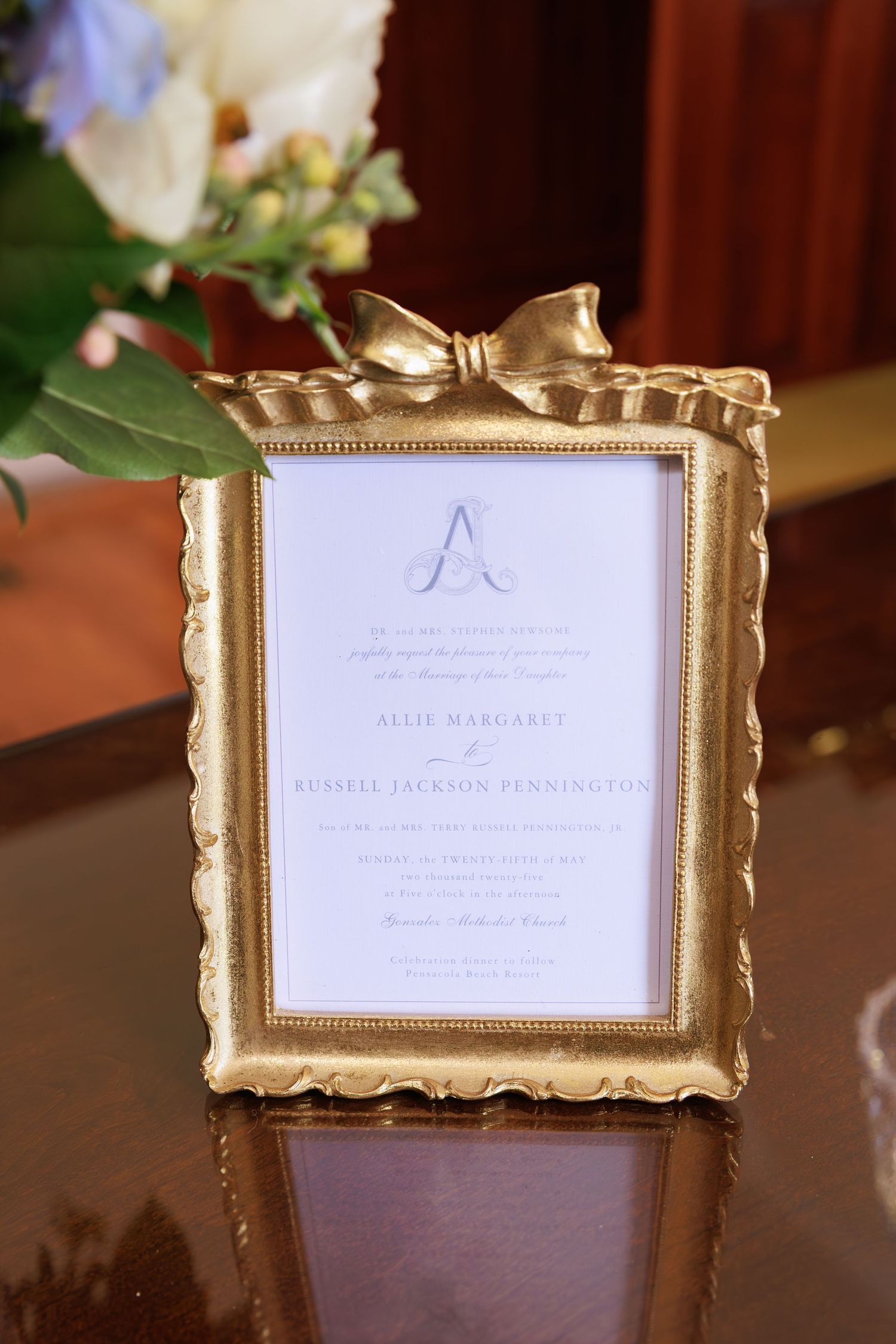 Elegant gold frame with bow displaying a wedding invitation on a polished wooden surface beside flowers.