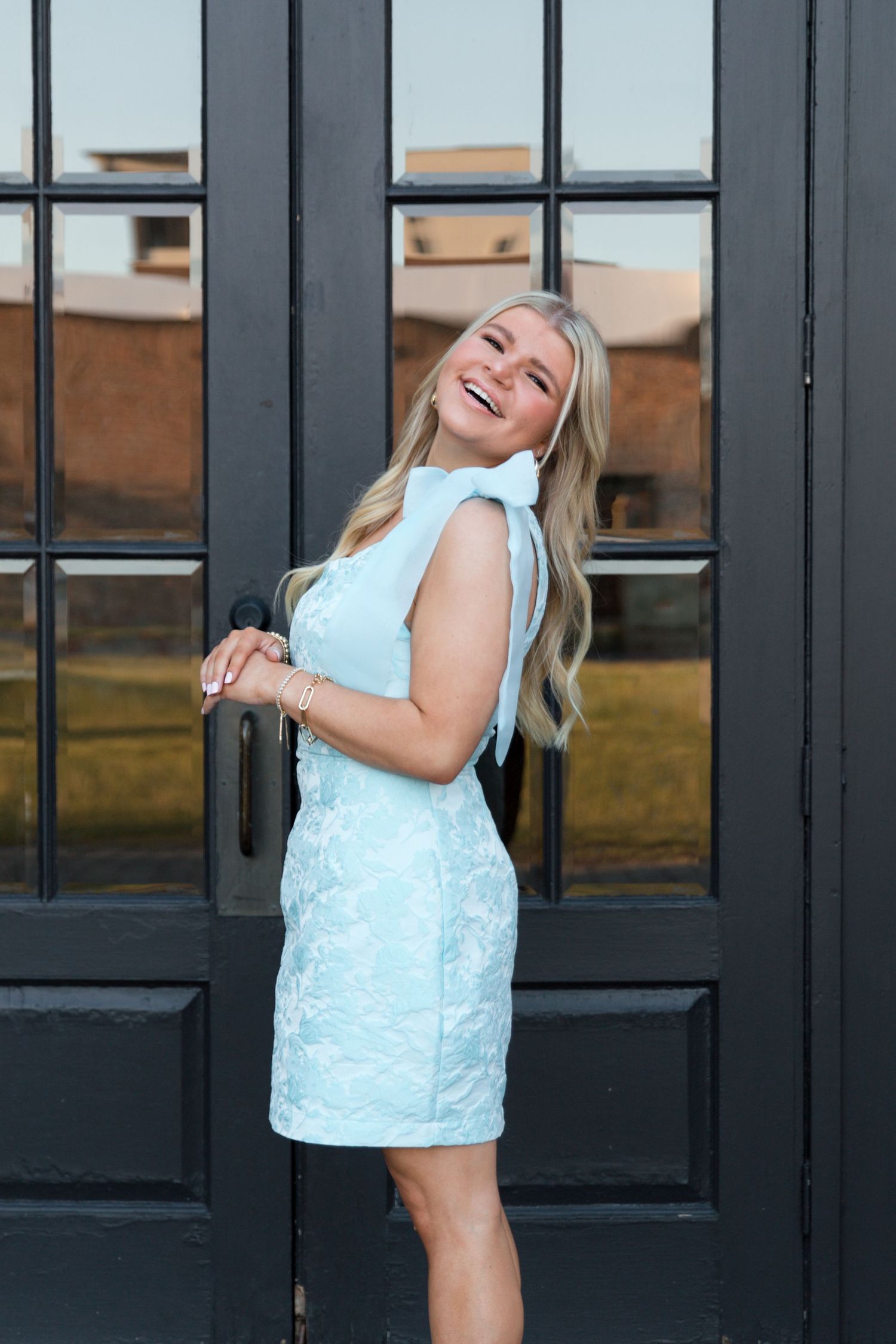 A person wearing a white lace dress poses playfully against dark French doors.