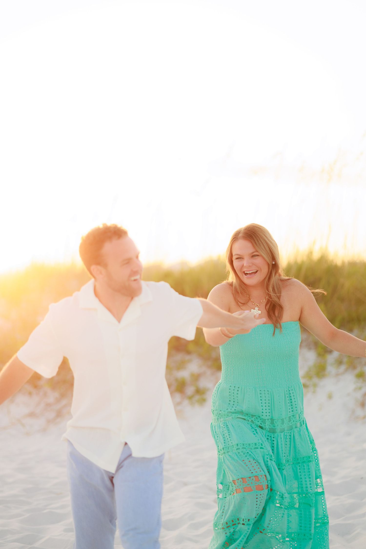 Playful couple laughing and running on sandy beach at sunset in summer attire.