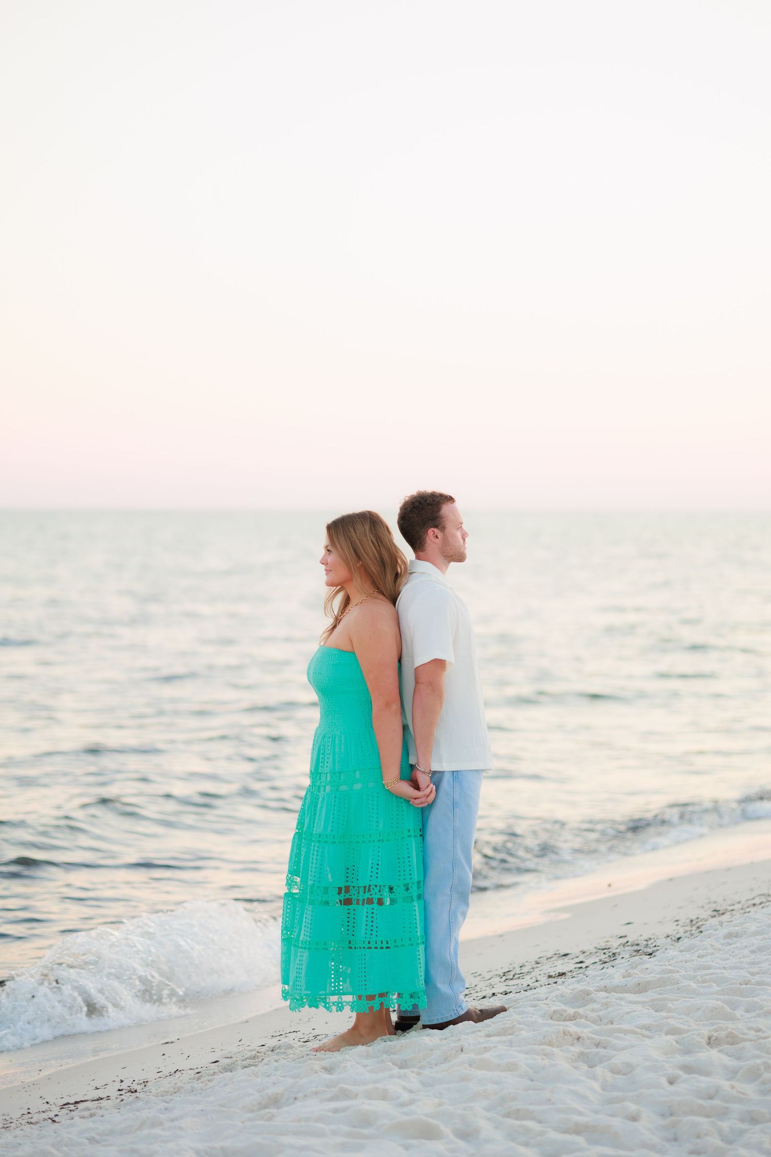 Romantic couple sits together on white sandy beach during sunset with gentle waves in background.