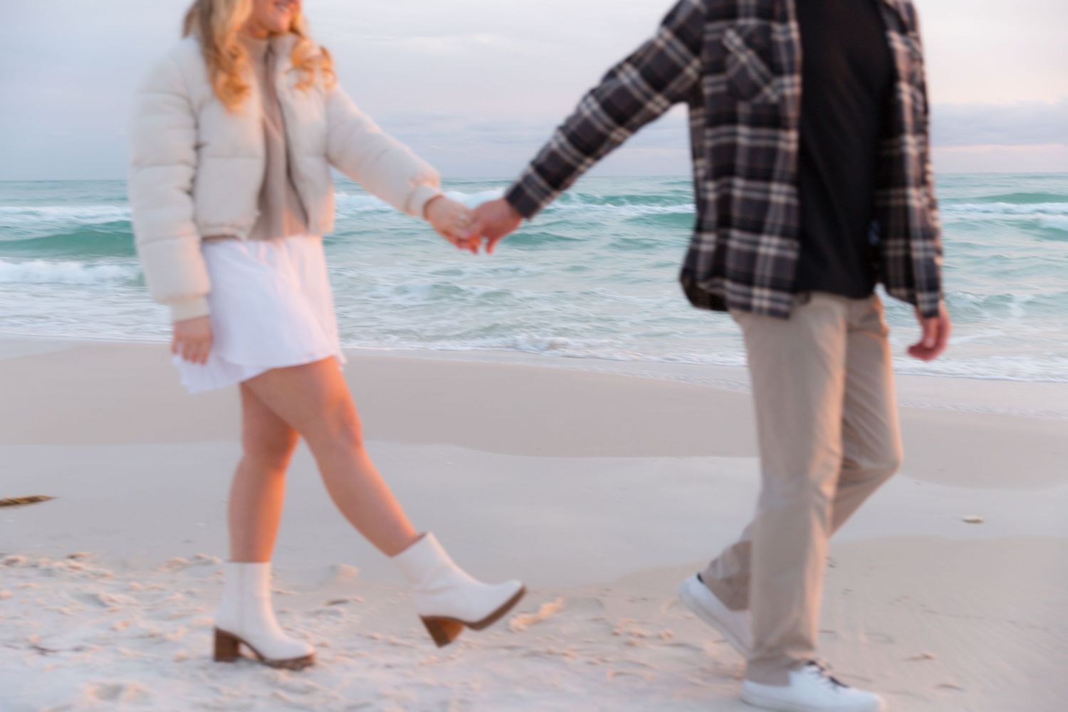 Two people holding hands while walking on a sandy beach by the ocean wearing casual clothes and white shoes.