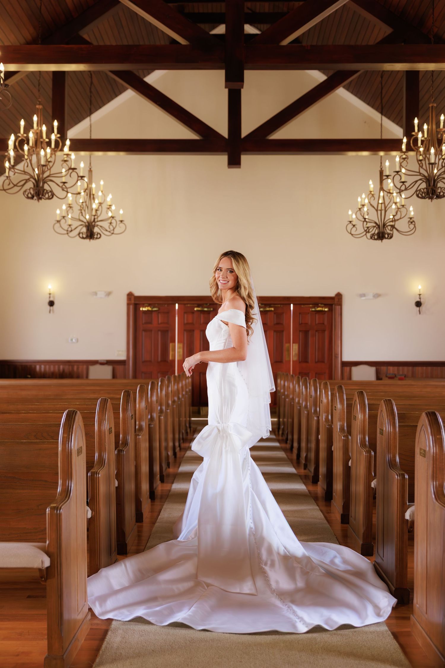 A flowing white wedding dress twirls gracefully between wooden church pews beneath elegant chandeliers.