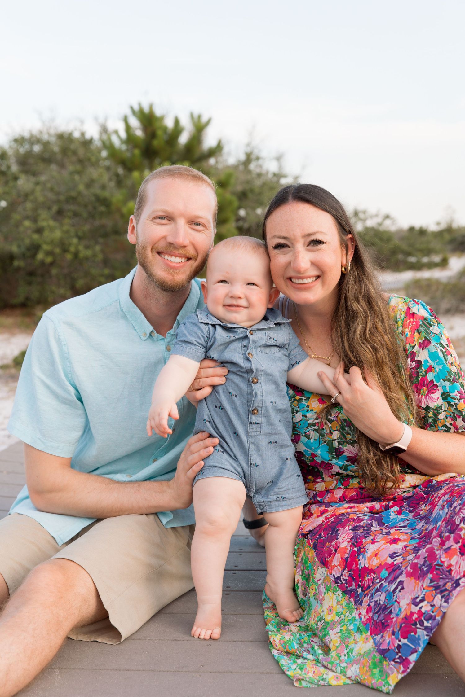 Family of three poses together on beach boardwalk wearing coordinating blue and floral outfits in sunset light.