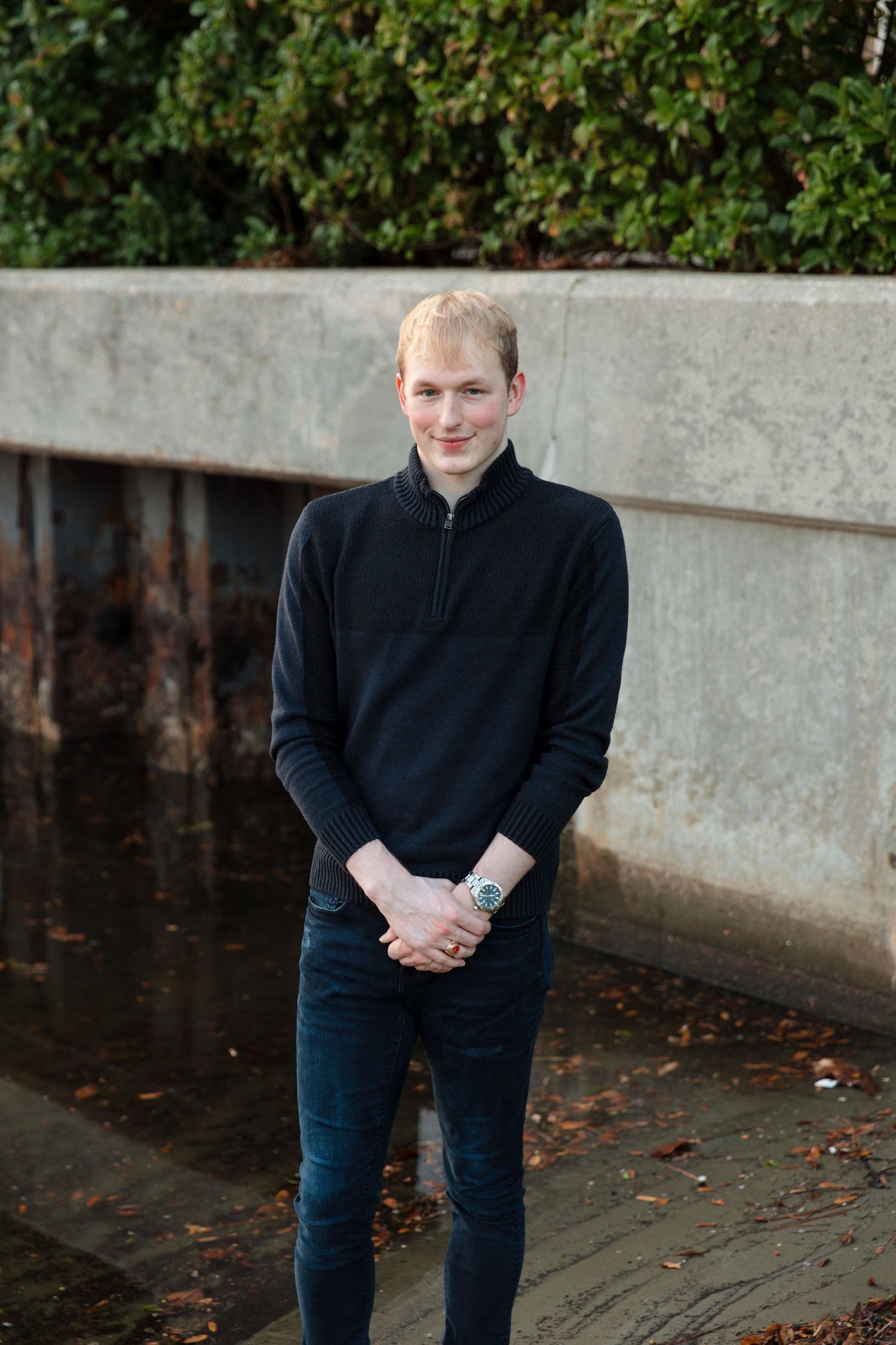 Young blond man in black zip-neck sweater and jeans standing outdoors near concrete wall with greenery.
