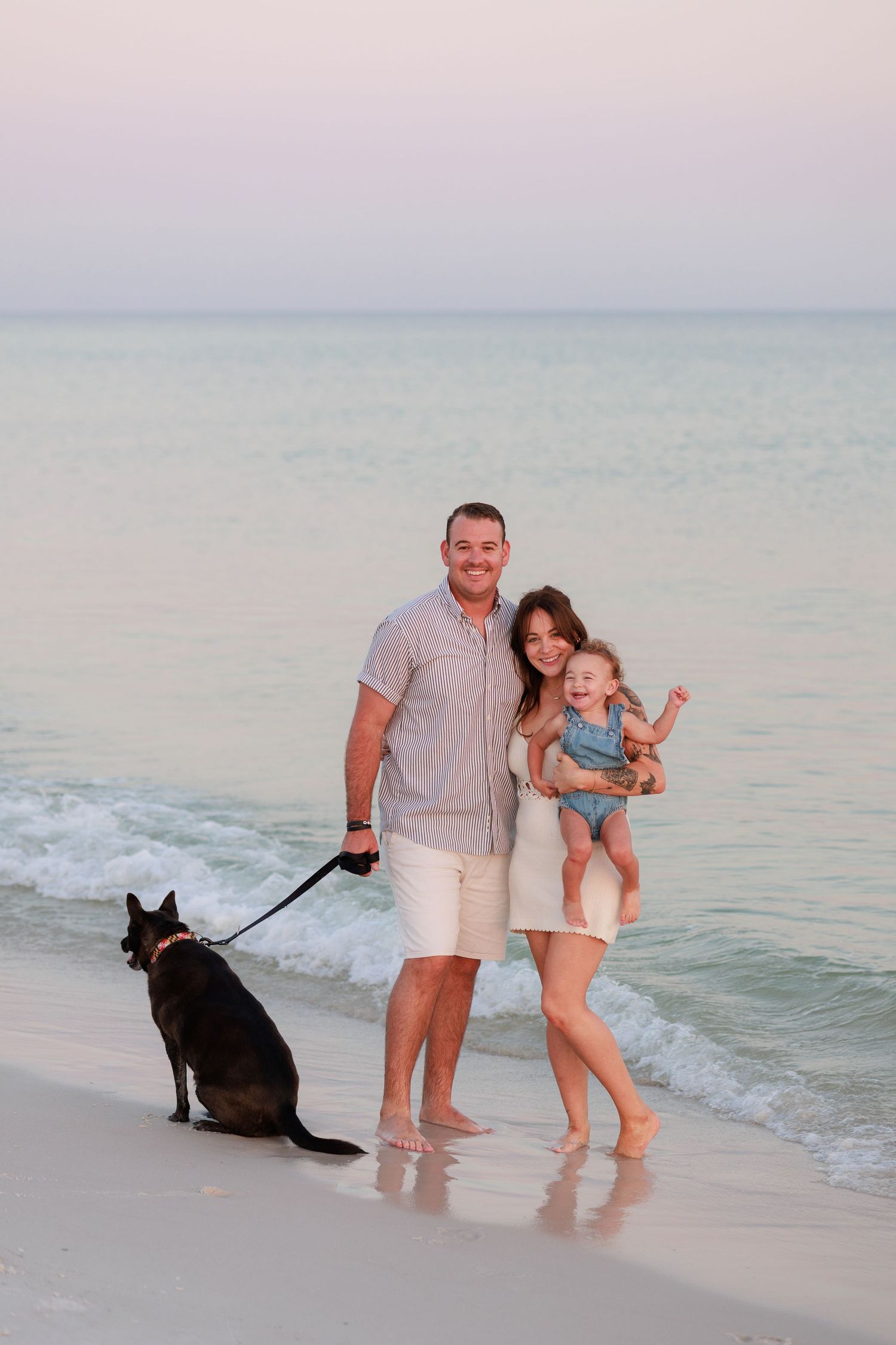 Family with dog posing on beach at sunset, waves in background.