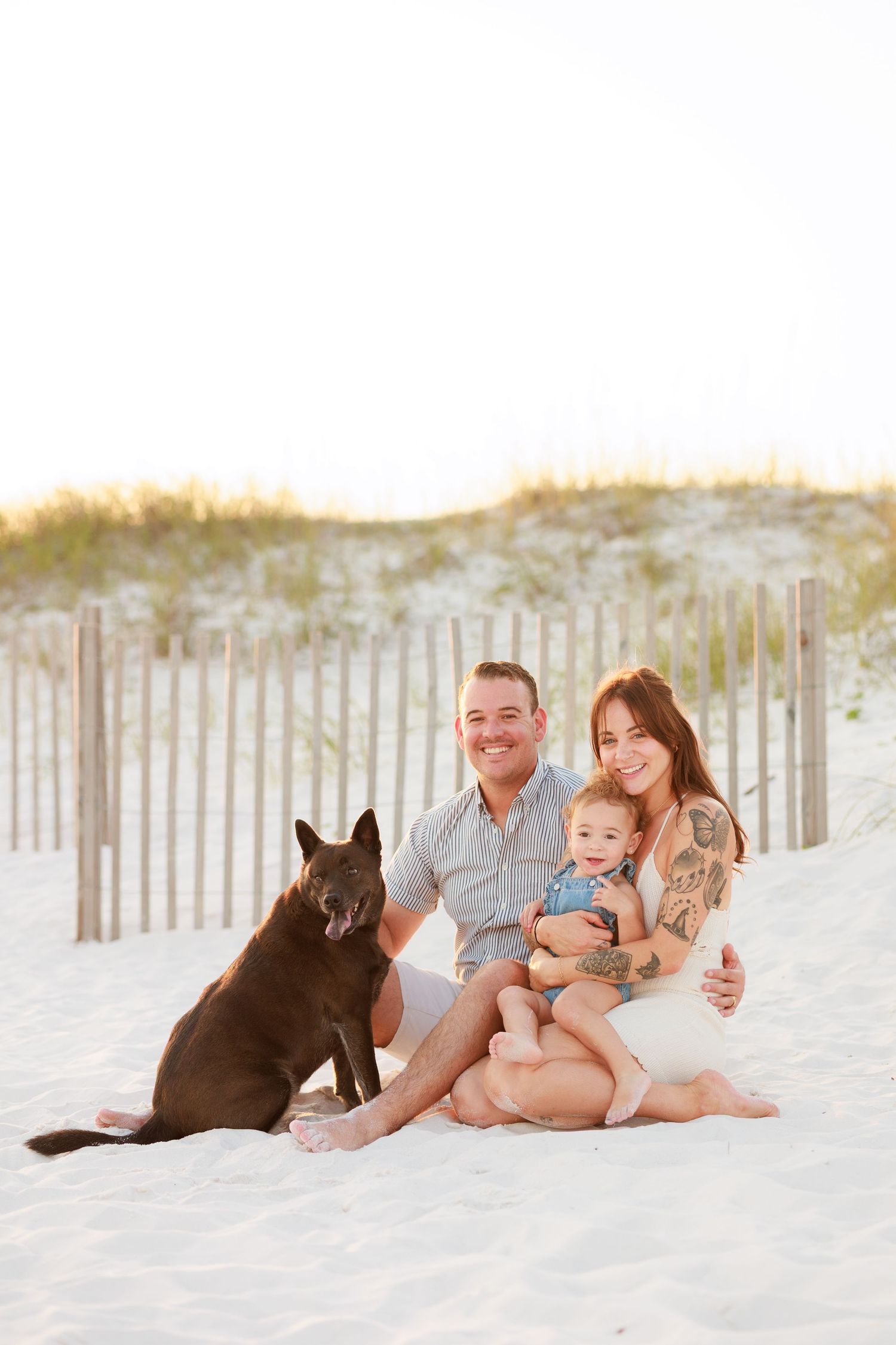 Family with baby and dark German Shepherd dog sitting on white sand beach at golden hour near dune fence.