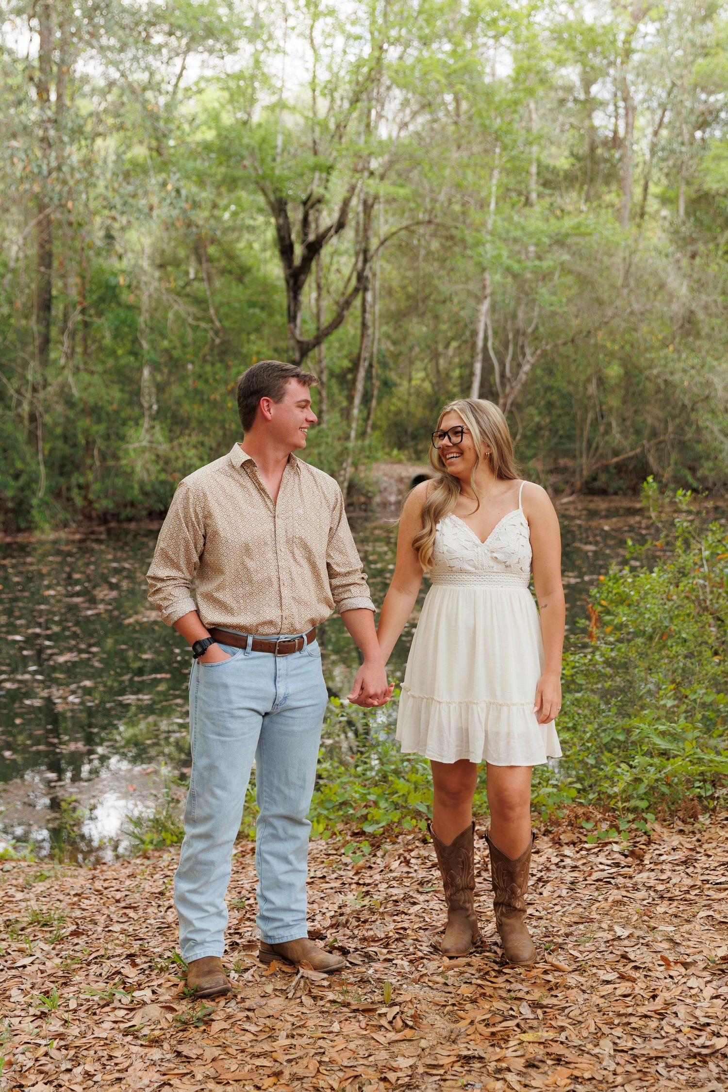 Couple holding hands and gazing at each other near a forest pond, dressed in country-style casual outfits.