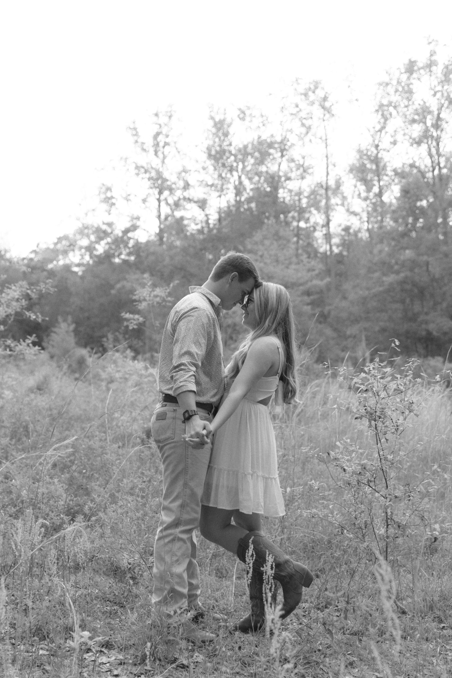 Black and white photo of a romantic couple nearly kissing in a sunlit meadow surrounded by tall grass.