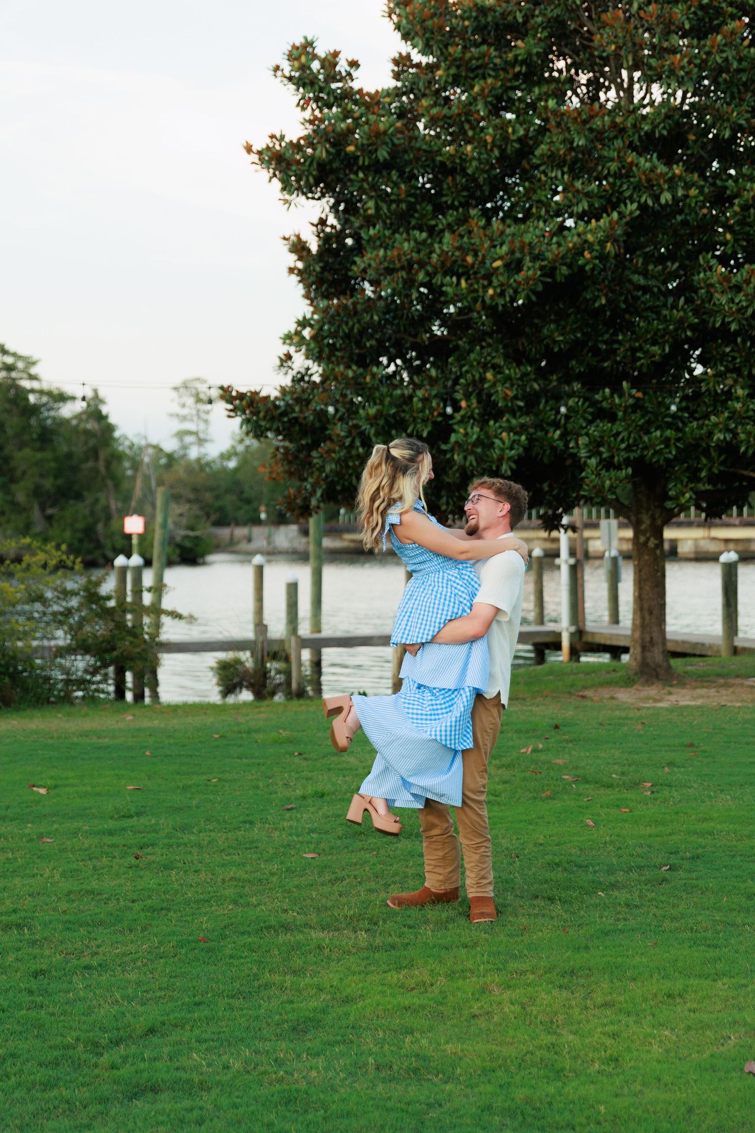 Joyful man lifting woman in a blue gingham dress on a green lawn near a waterfront at dusk.