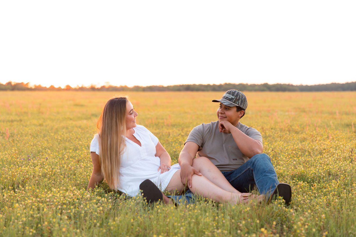 Couple sitting together in a sunny golden field of wildflowers, sharing a romantic moment at sunset.