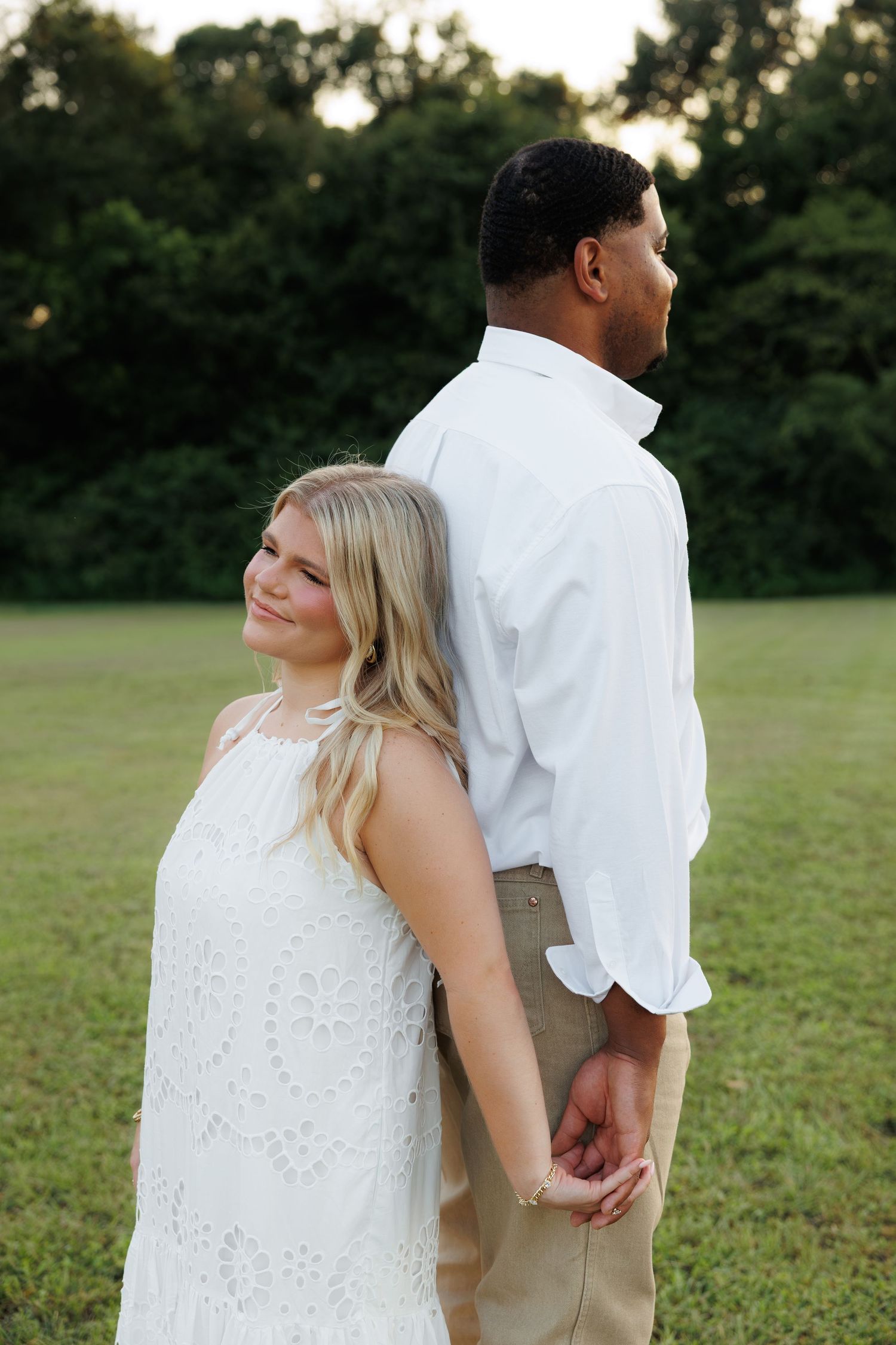A couple in white outfits holds hands back-to-back in a green field, the woman smiling warmly at the camera.
