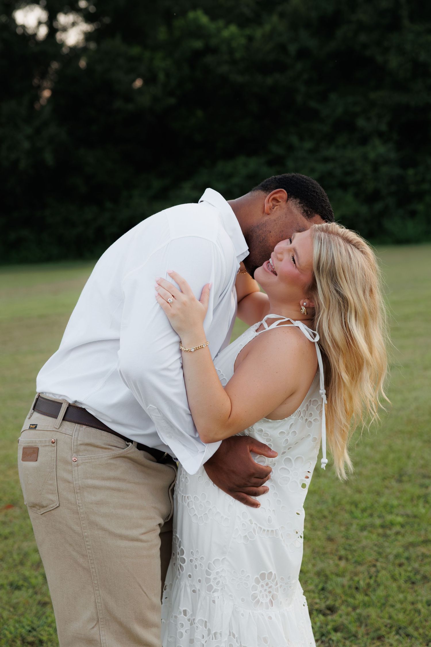 Romantic interracial couple embracing and kissing outdoors in a green field, wearing coordinated white and khaki outfits.