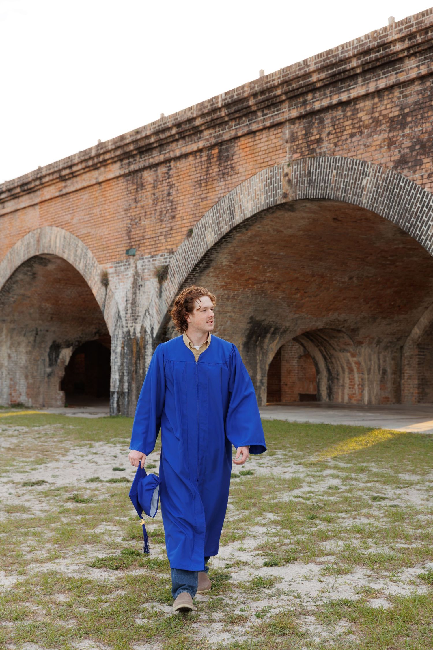 Graduate in blue cap and gown walking near historic brick archway.