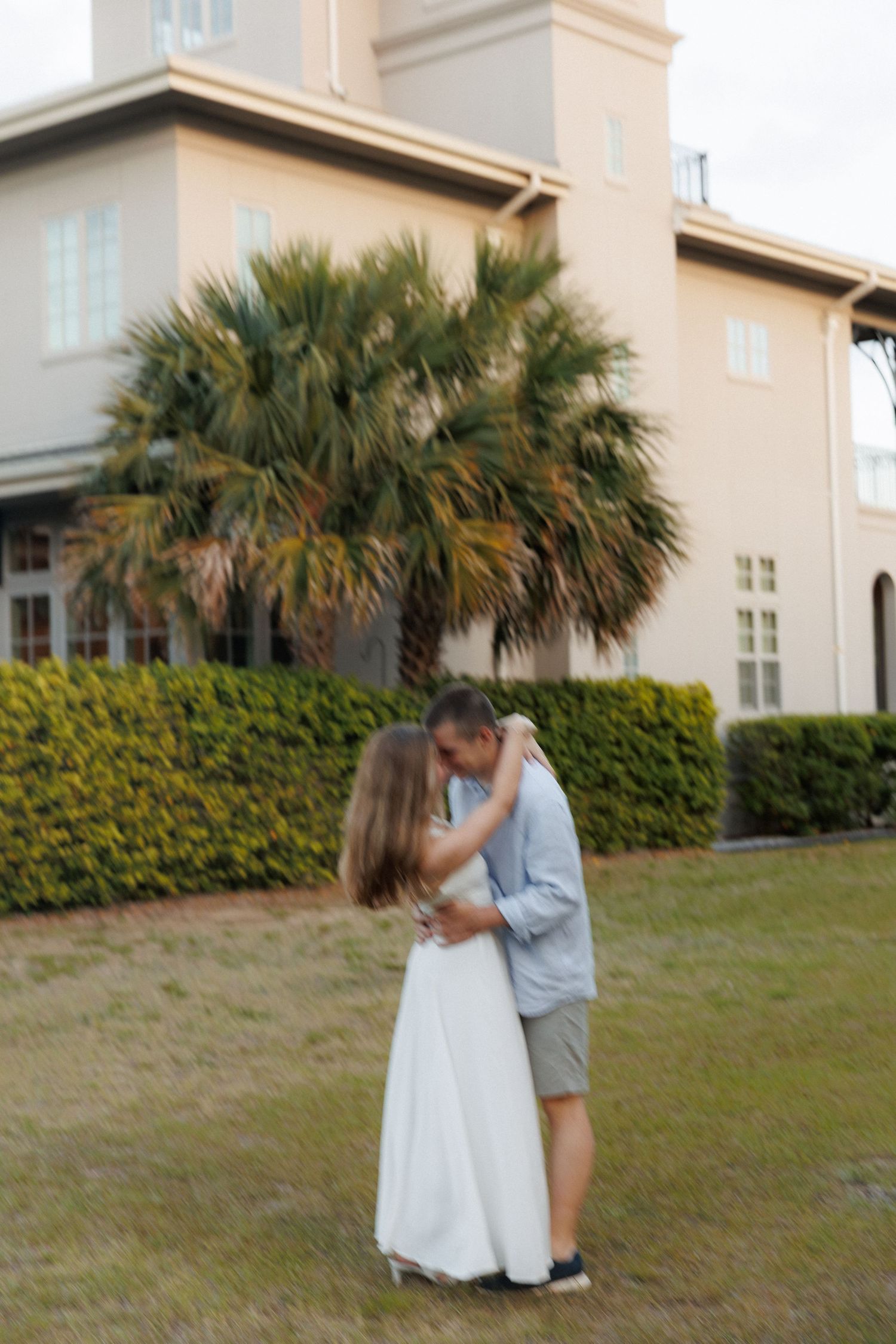 Couple sharing a tender kiss on a lawn with palm trees and a large building in the background.
