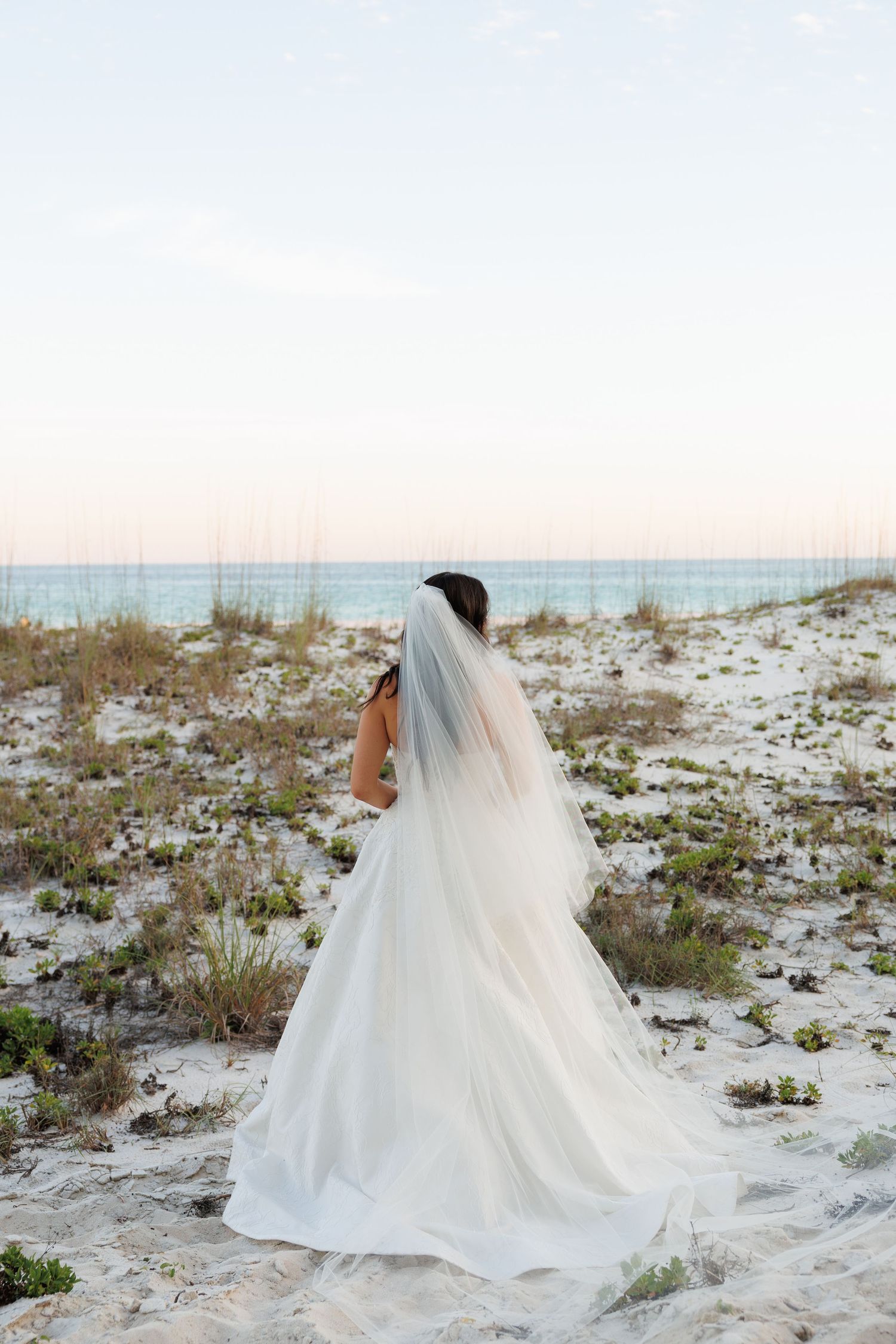 Bride in white gown with flowing cathedral veil walks along white sand beach at dusk.