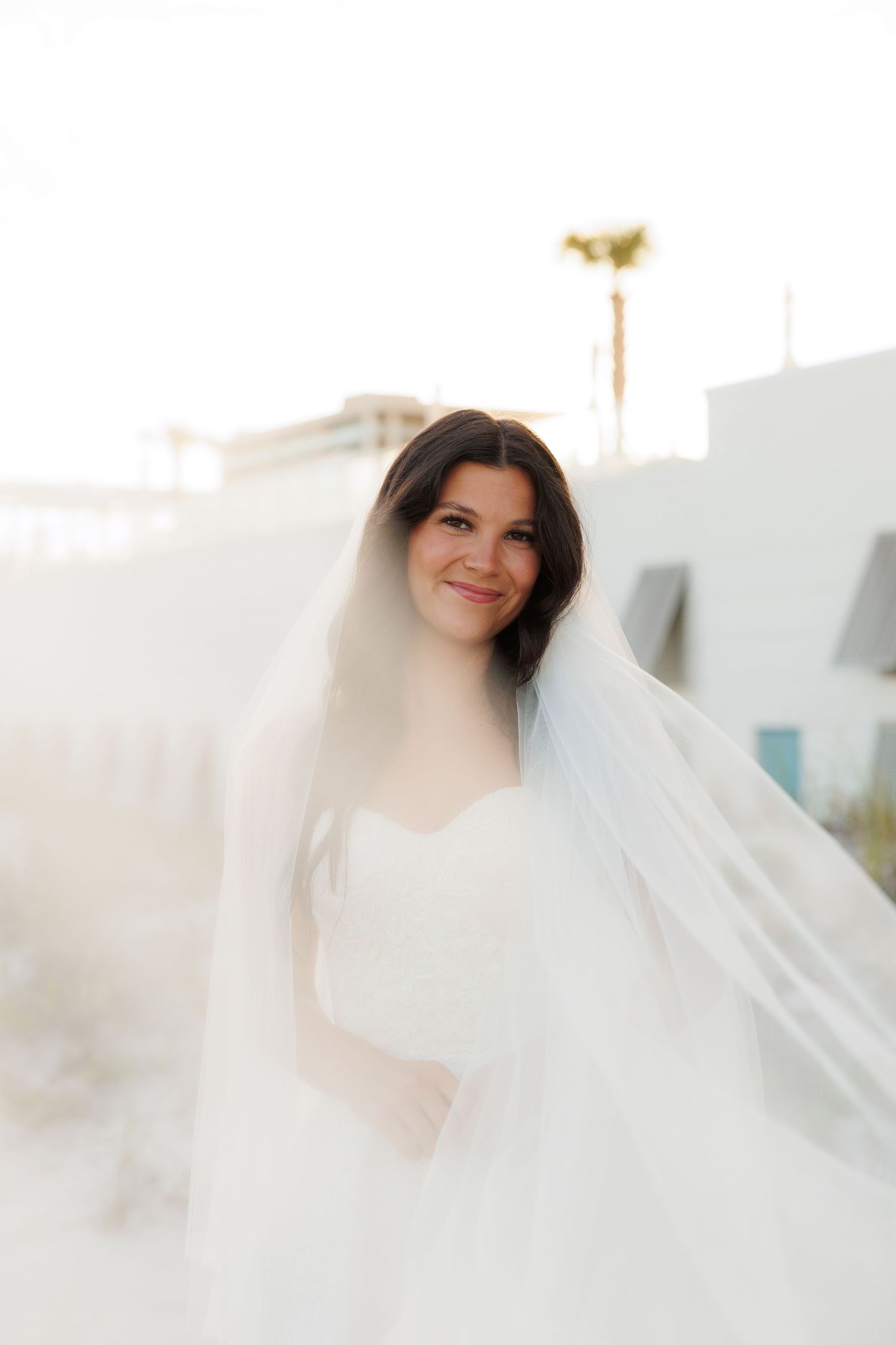 Smiling bride in white strapless gown and flowing veil poses outdoors with soft golden backlight.