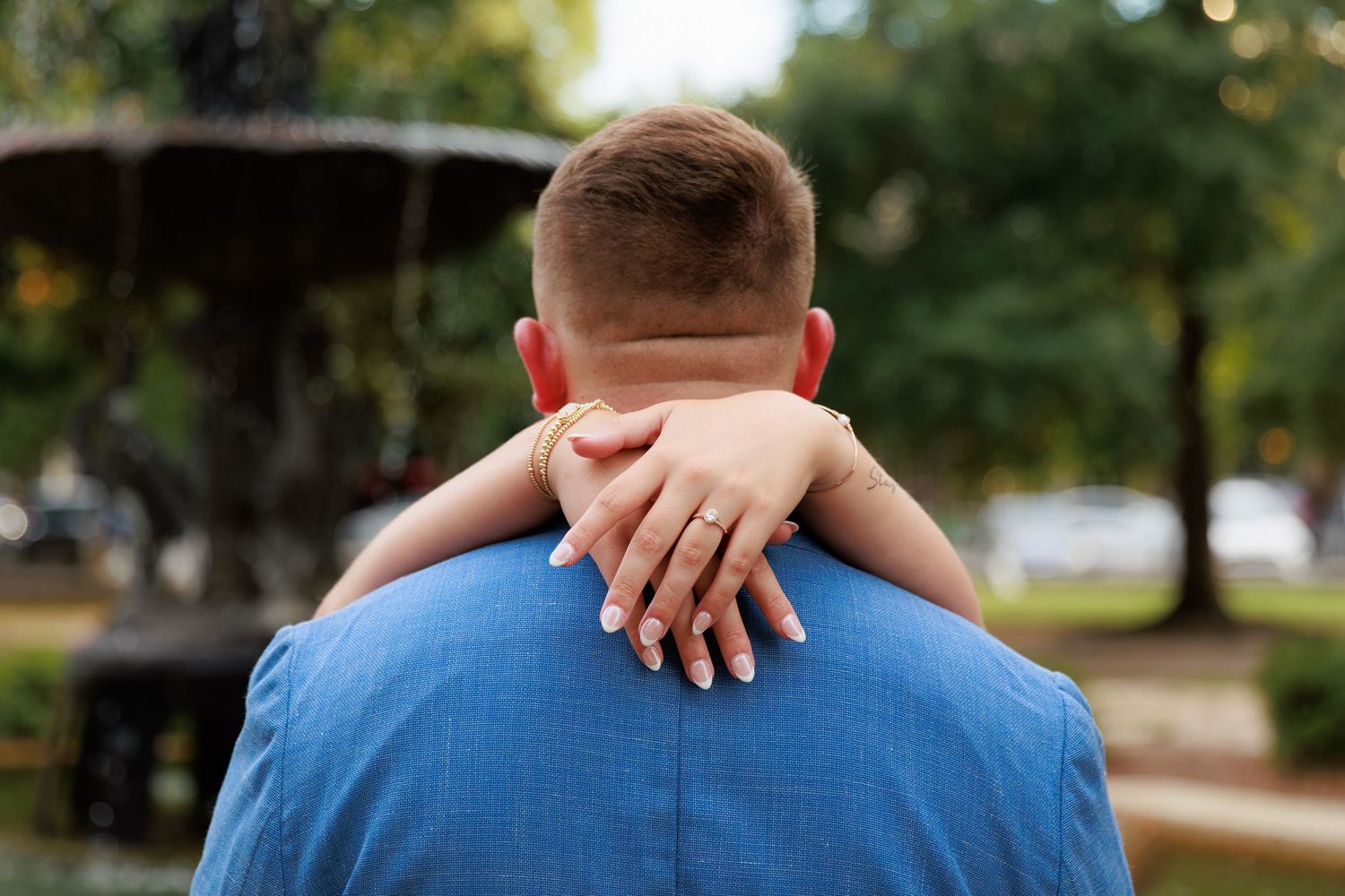 Couple embracing near a fountain in a park, woman's hand with engagement ring resting on man's shoulder.