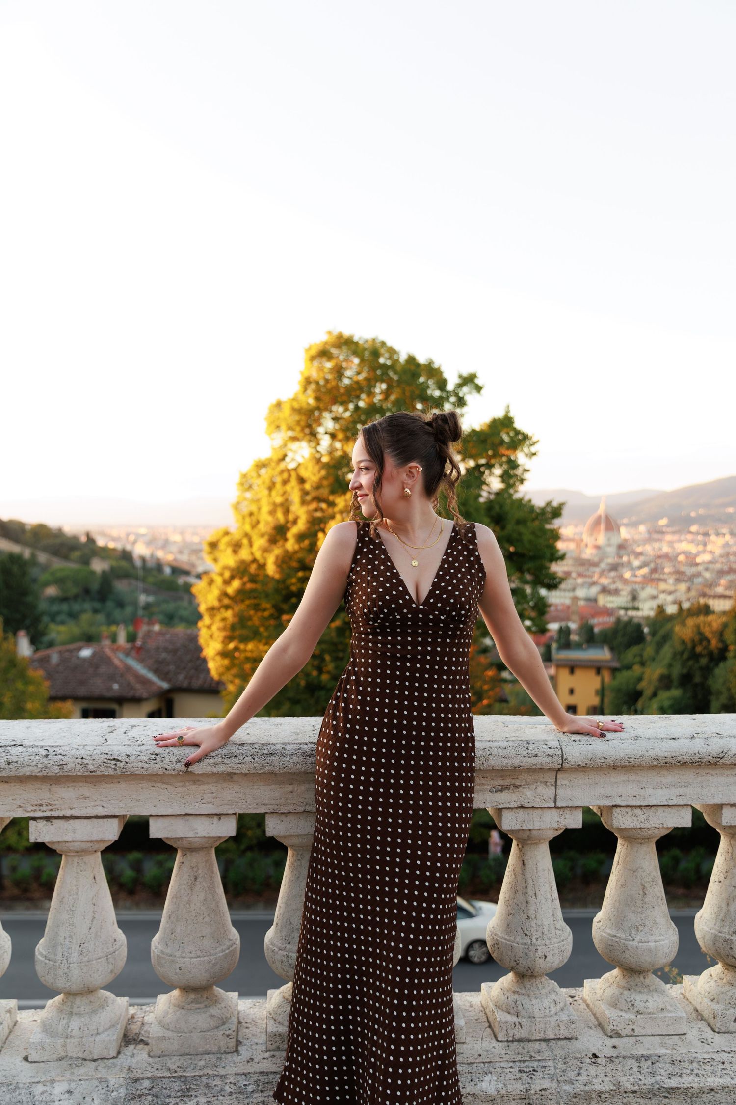 Elegant woman in brown polka dot maxi dress posing at stone balustrade with Florence cityscape behind.