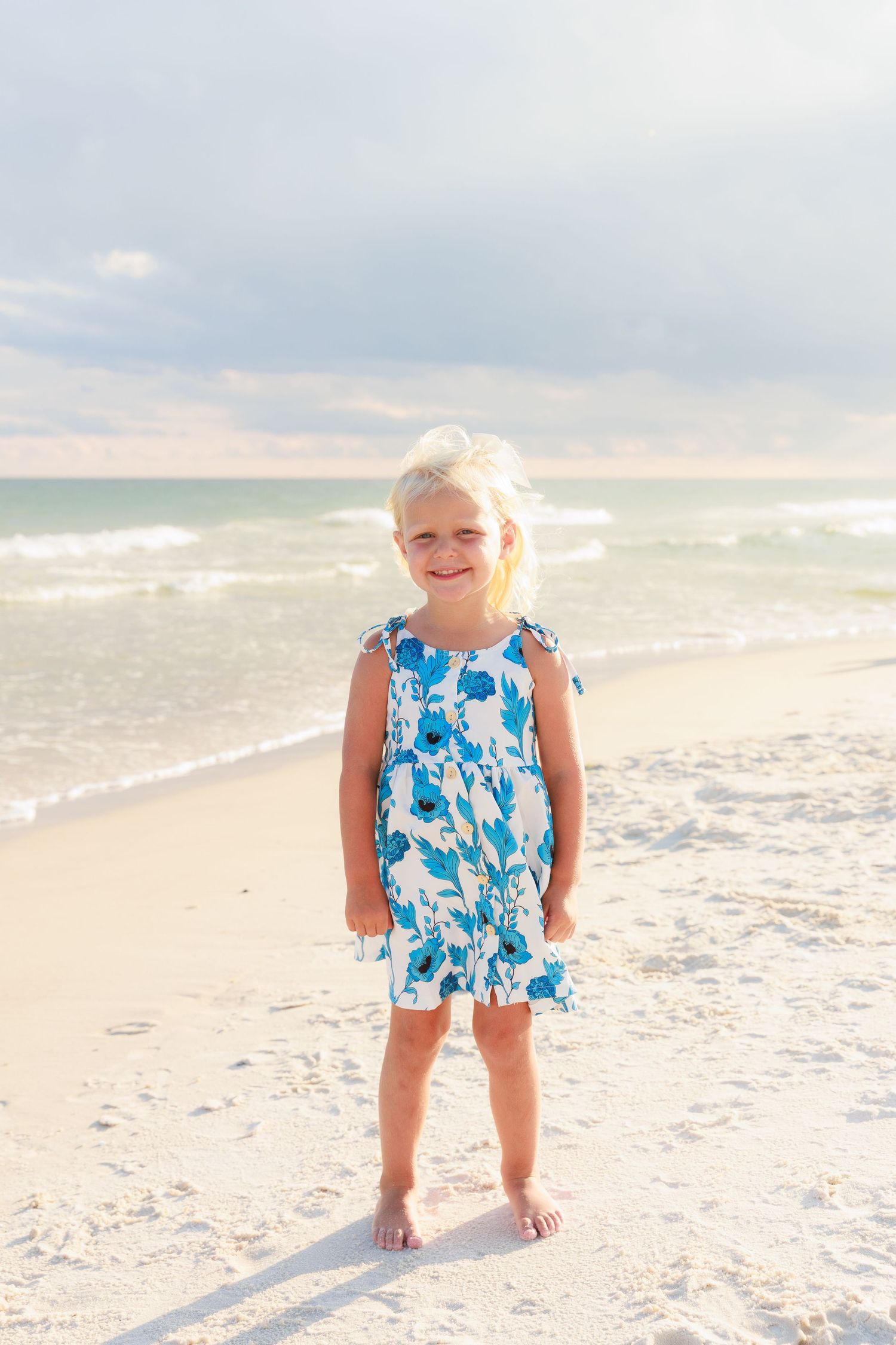 Smiling blonde girl in blue floral dress standing barefoot on sandy beach with ocean waves behind her.