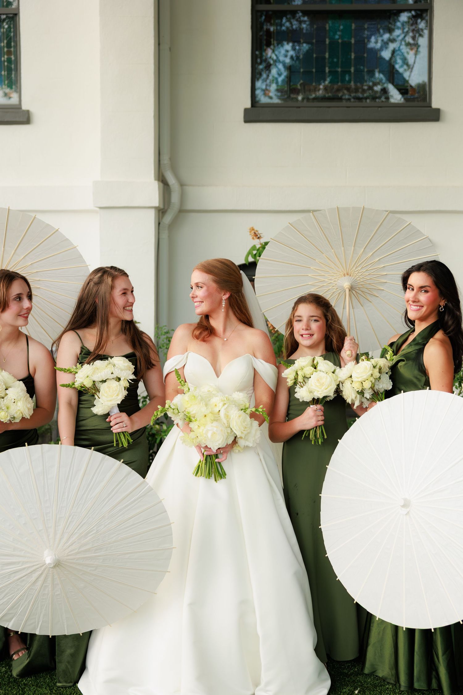 Bride in white gown with bridesmaids in olive green dresses holding white parasols and bouquets outdoors.