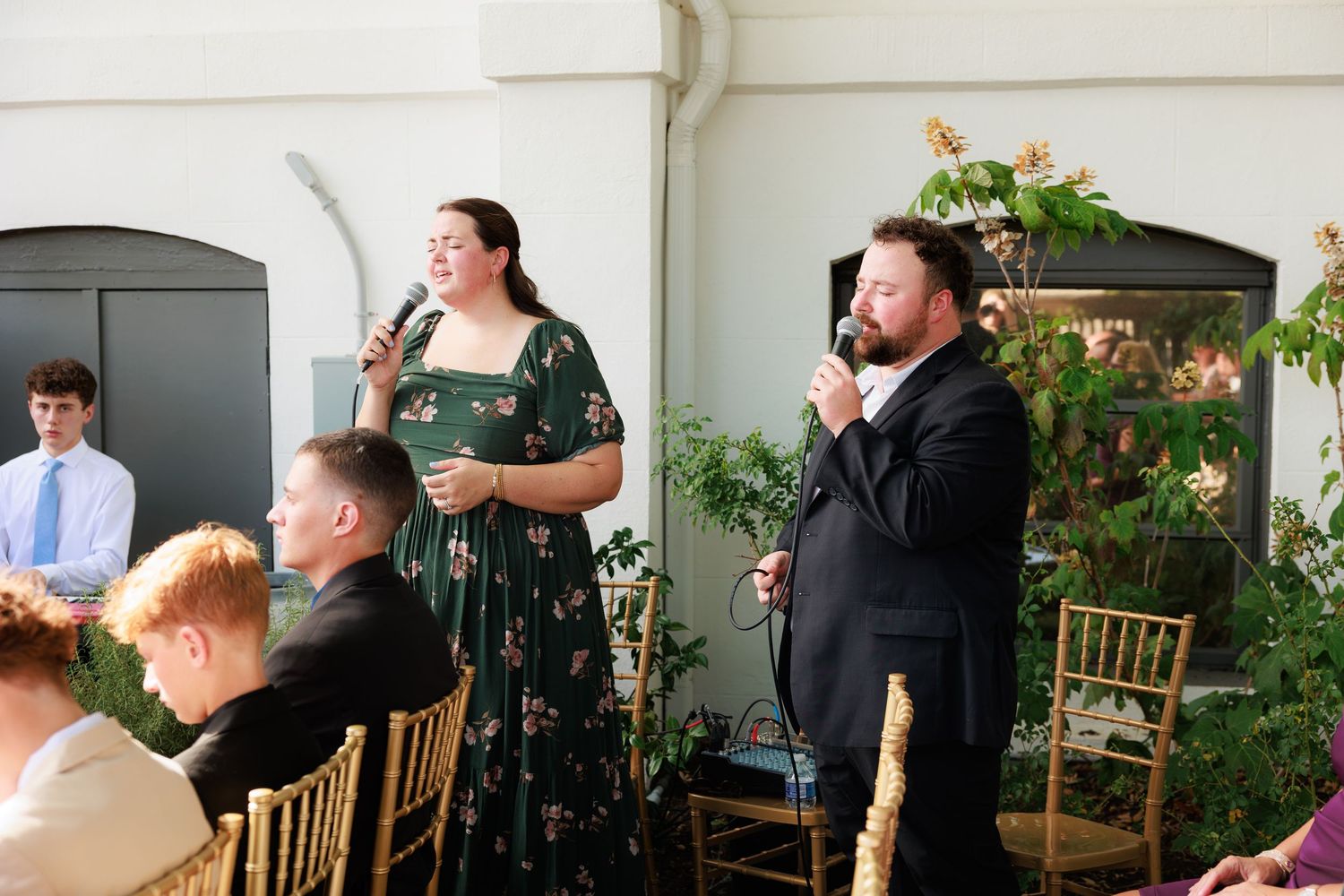 A man and woman in formal attire speak at microphones during an elegant wedding reception indoors.