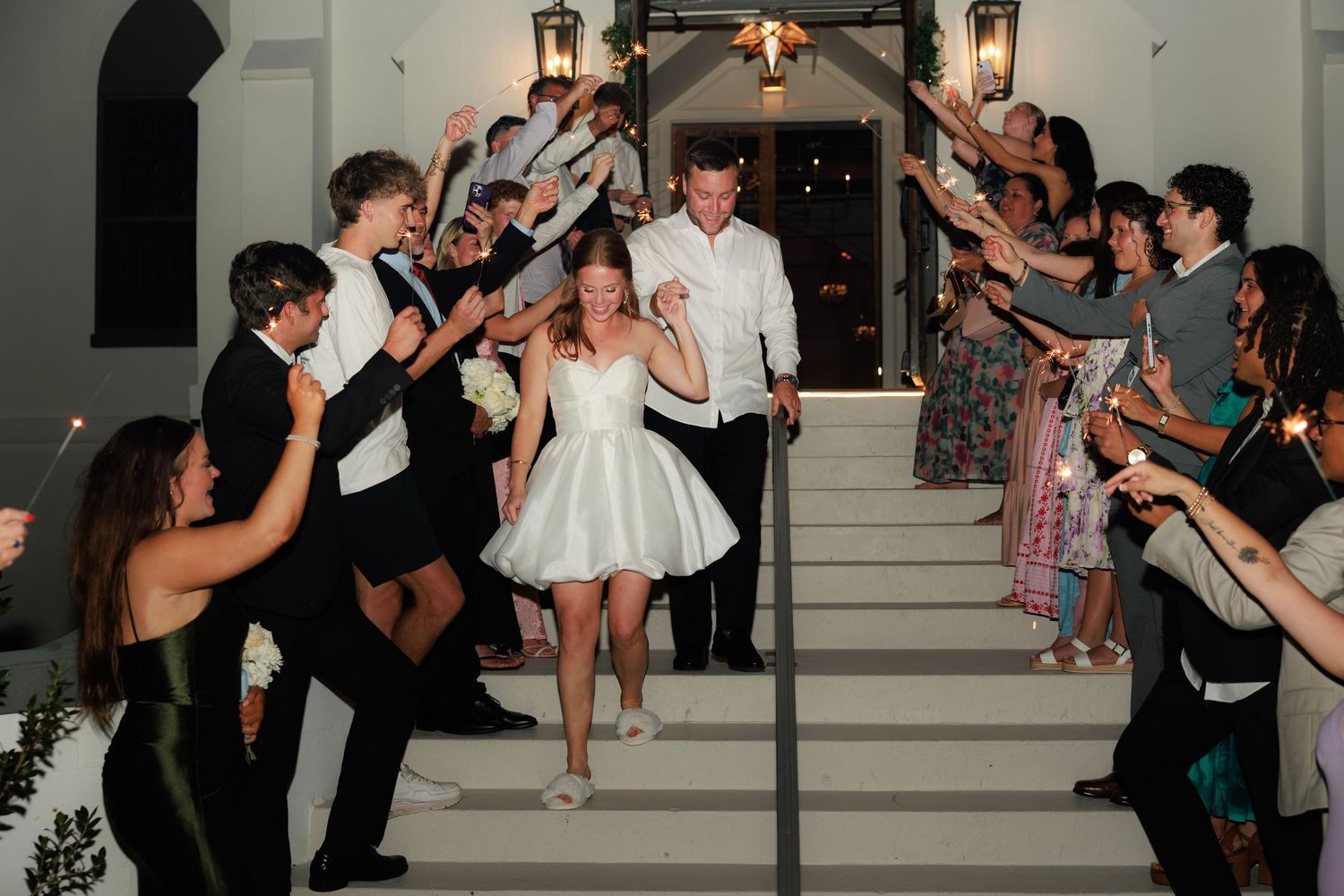 Bride and groom exit wedding venue on steps surrounded by cheering guests holding sparklers at night reception.