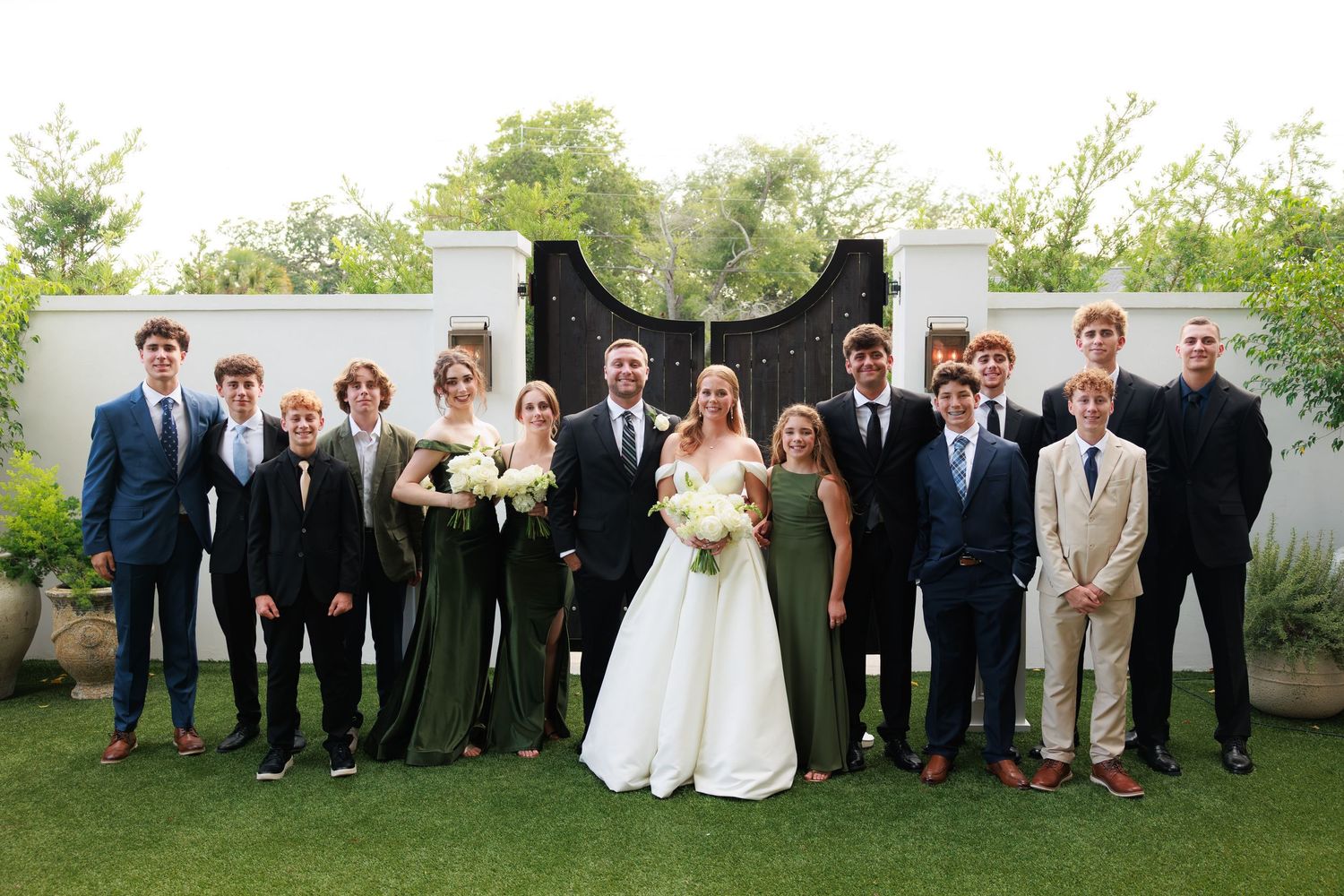 Bride and groom pose with wedding party in olive green dresses and navy suits before ornate black gate.