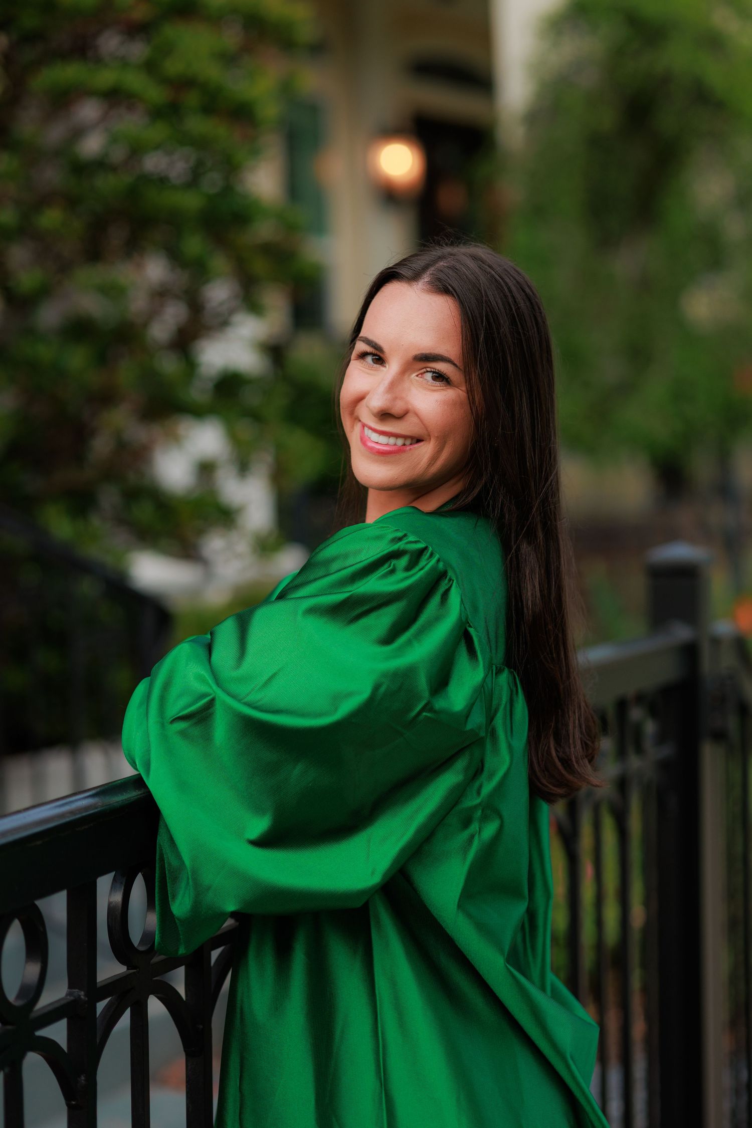 Young woman in green graduation gown smiling on outdoor balcony with lush greenery background.