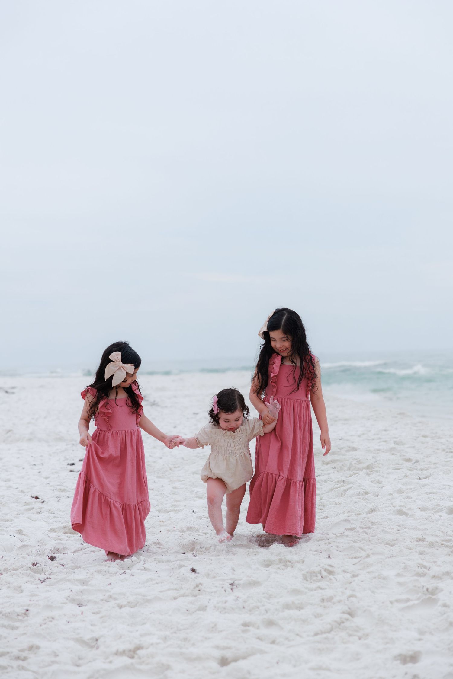 Three girls in pink dresses hold hands on a white sandy beach with ocean waves in the background.