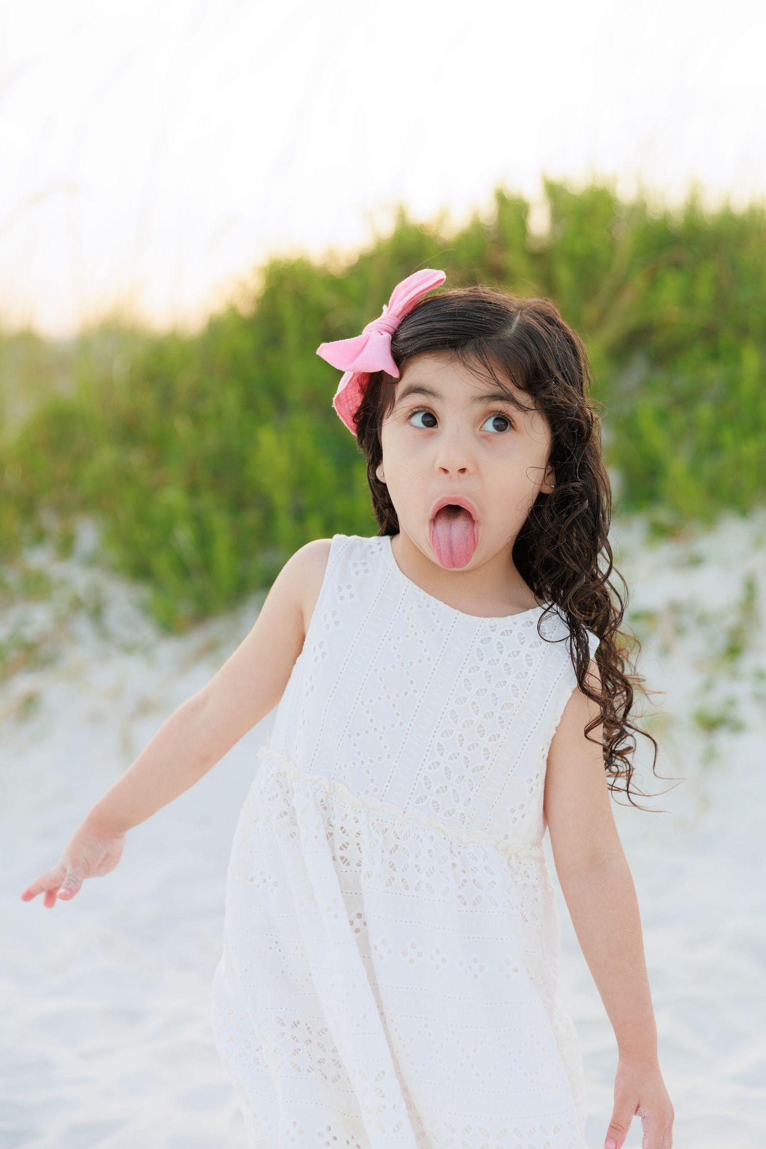 A young girl in a white dress with a pink bow sticks out her tongue playfully on a sandy beach.