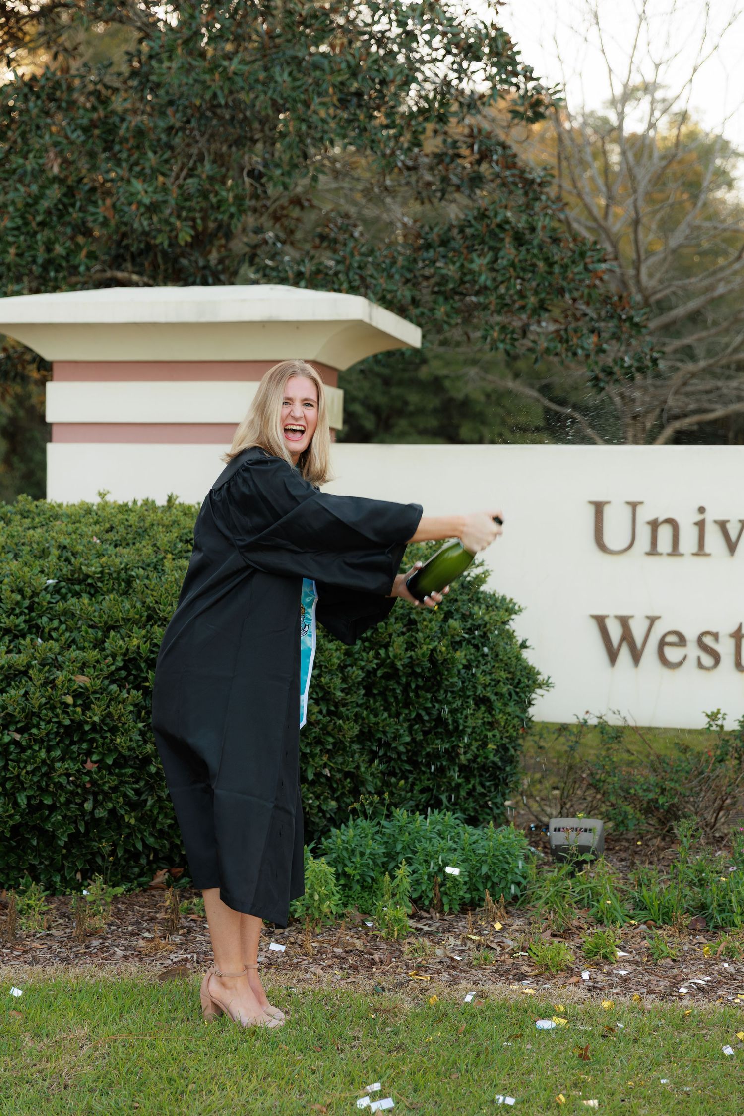 Graduate in black cap and gown poses playfully by university entrance sign during graduation photoshoot.