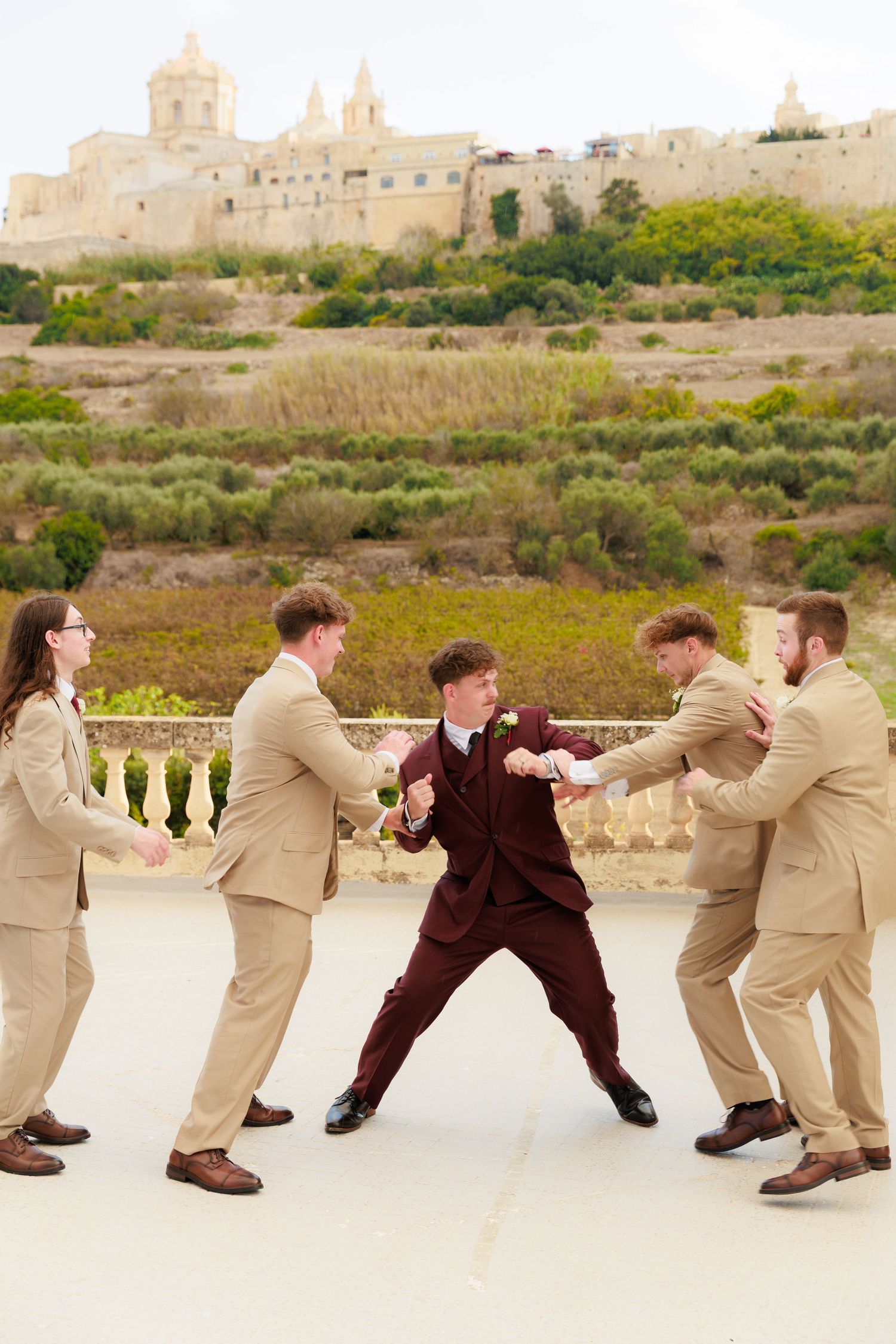 A playful wedding party poses in beige and burgundy suits with a historic castle backdrop in Malta.