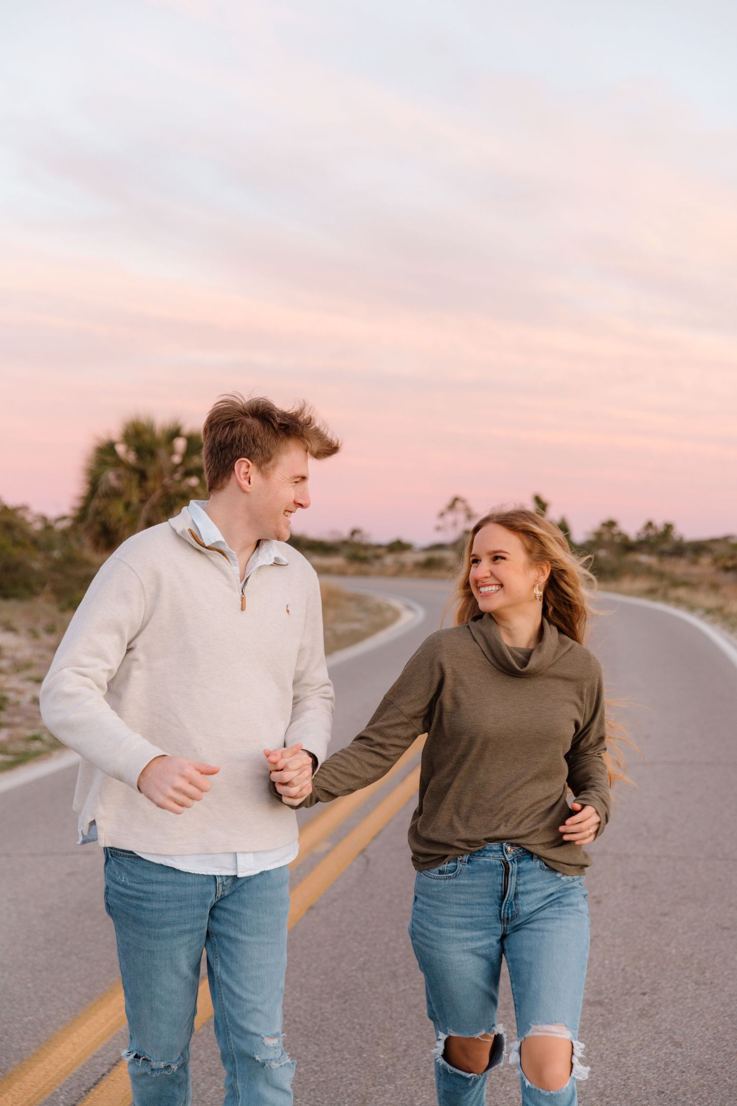A couple wearing jeans share playful moments walking along an empty road at sunset.