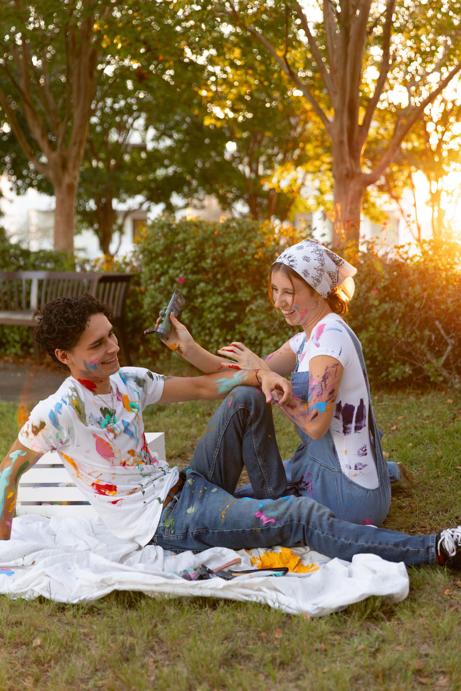 Two people in paint-splattered clothes sit on a blanket having a picnic at sunset in a grassy park.